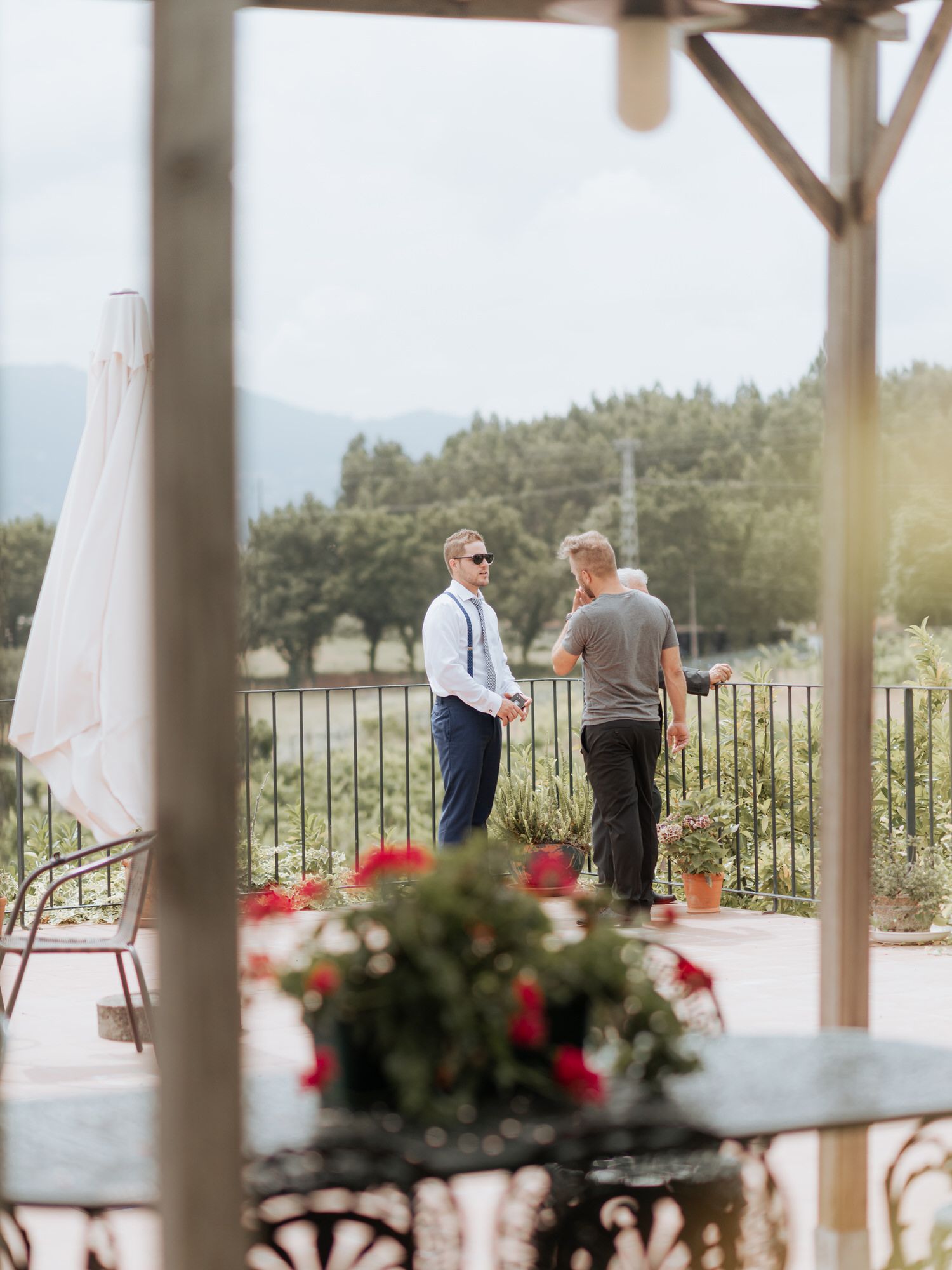 Two people conversing on a patio with red flowers and mountain views in the background.