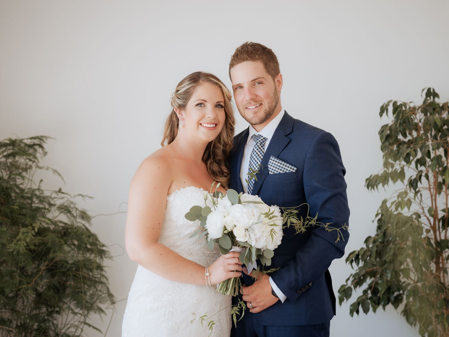Newlywed couple in elegant wedding attire poses with white floral bouquet against greenery backdrop.