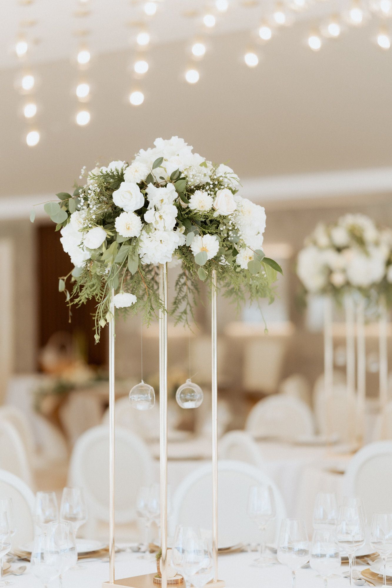 Elegant white floral centerpiece arrangement on tall modern metal stand with hanging glass ornaments at wedding reception.