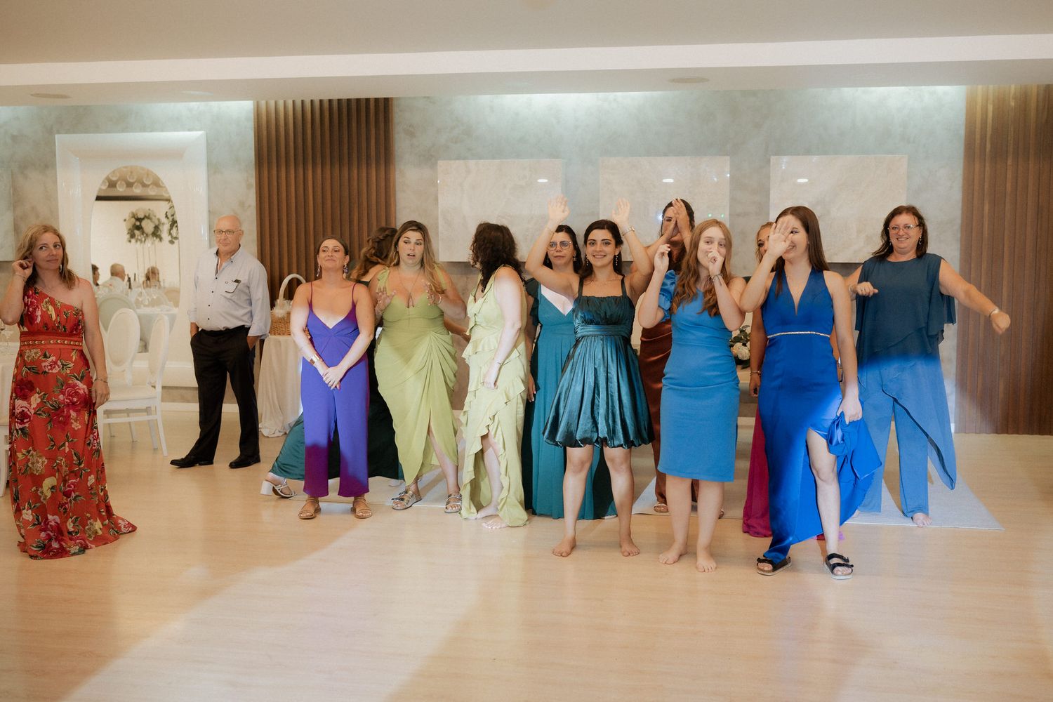 A group of women in colorful formal dresses stand together in a line at an elegant indoor venue during a celebration.