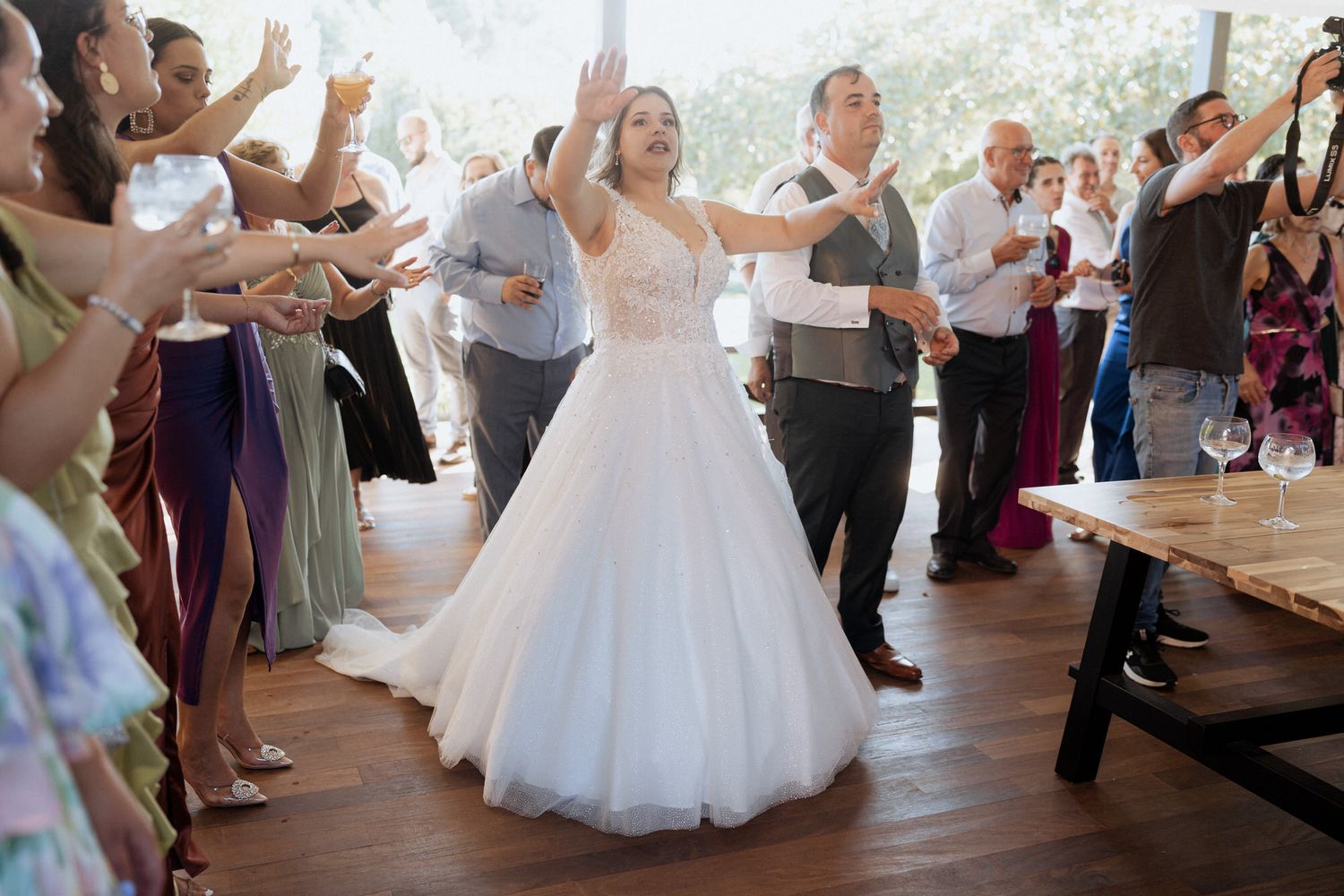 Wedding celebration with guests cheering and clapping as newlyweds dance joyfully in an indoor reception venue.