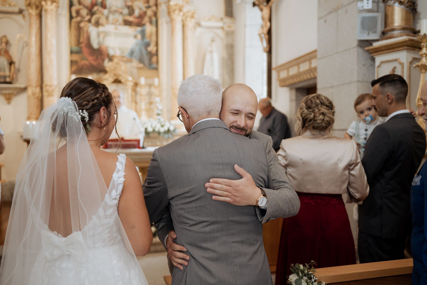 Inside an ornate church, two people embrace during a wedding ceremony while others stand nearby.