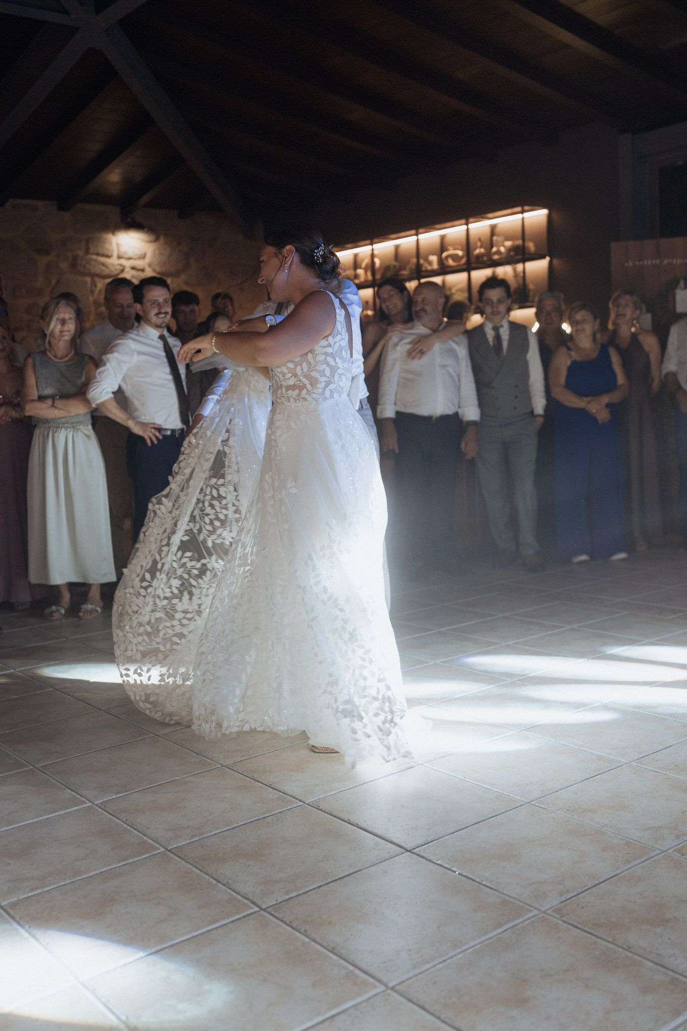 A couple shares their first dance as newlyweds on a tiled floor at a rustic reception venue with glowing lights above.