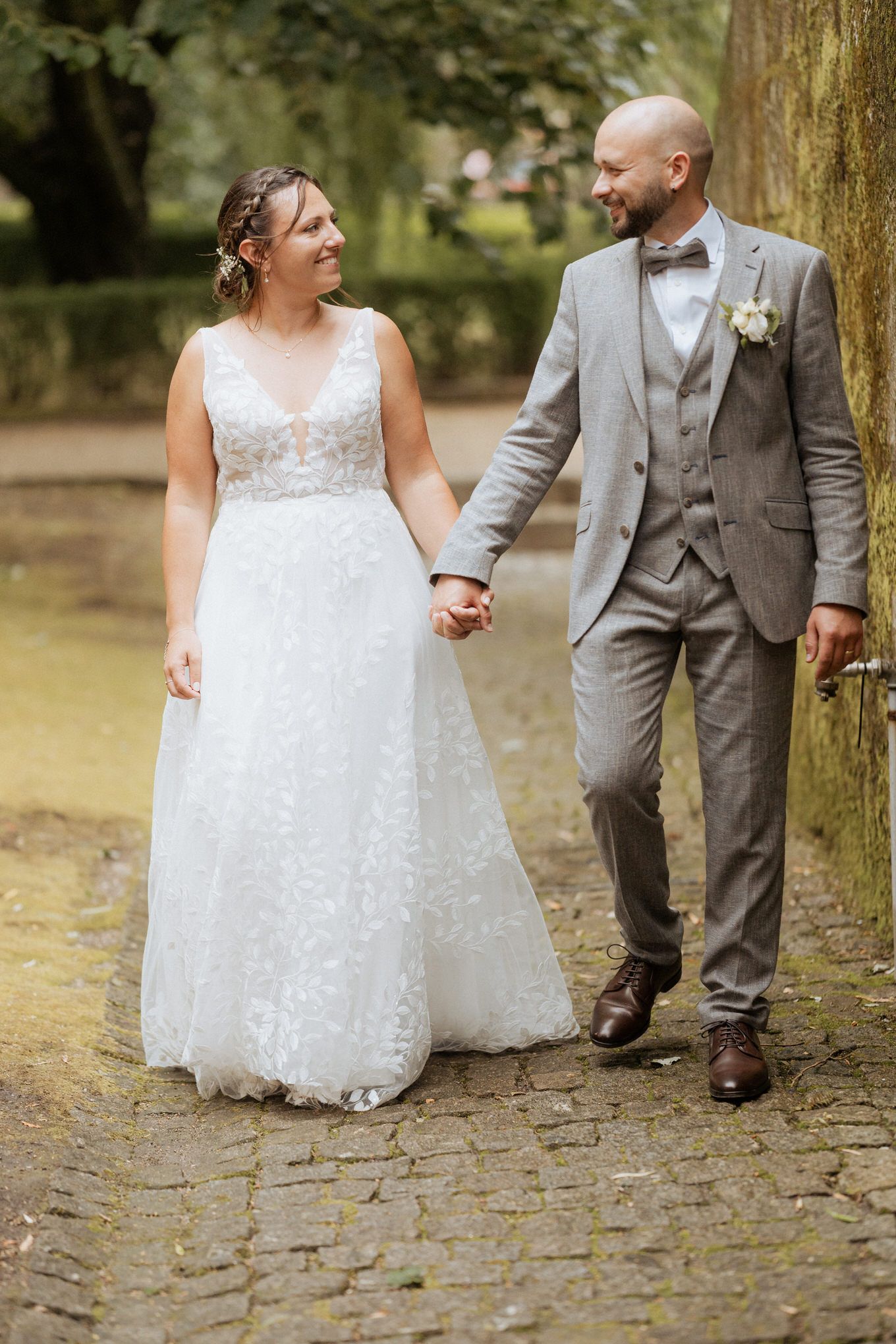 A newlywed couple holding hands while walking along a garden path, the bride in white lace and groom in grey suit.