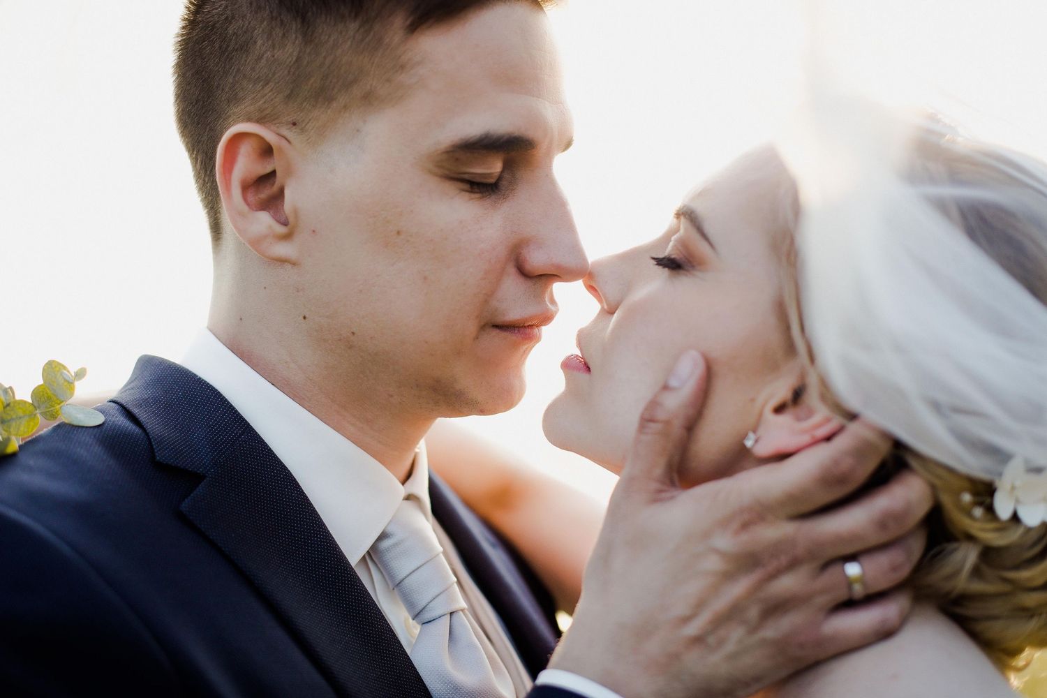 A groom in a navy suit tenderly holds his bride's face as they share an intimate moment before their first kiss.