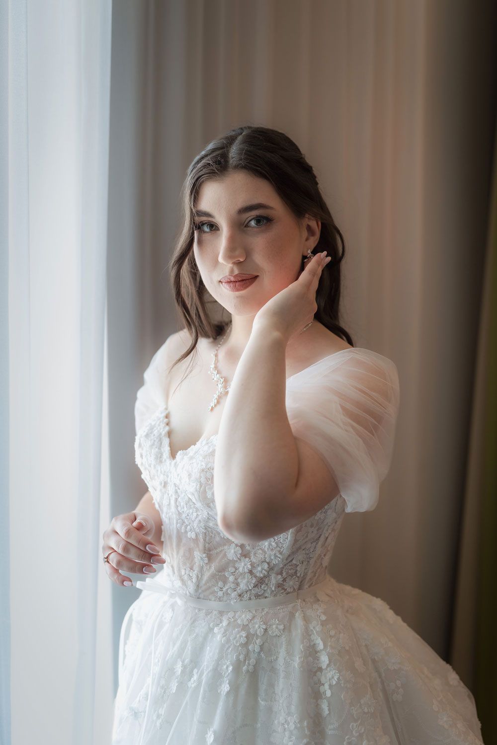 Elegant bride in white lace off-shoulder wedding gown with tulle sleeves posing gracefully by a window.