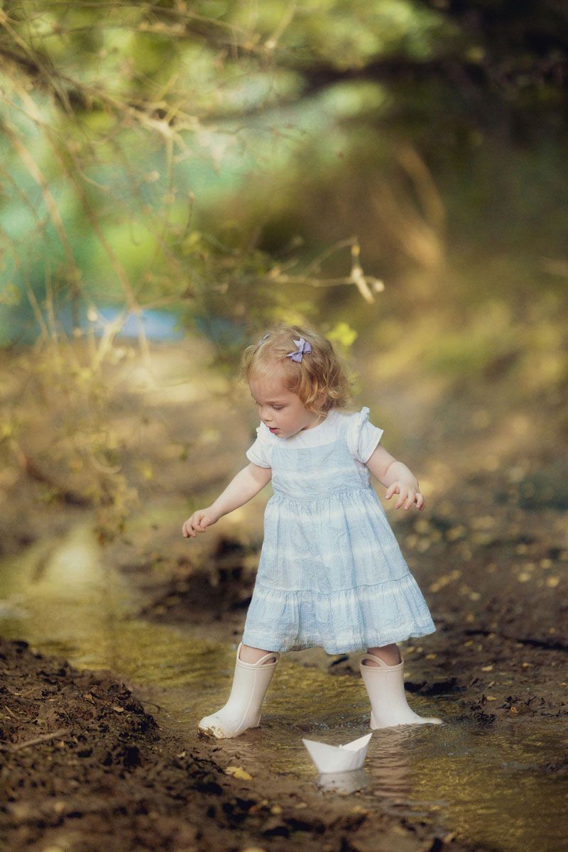 A little girl in a blue dress and white boots plays with a paper boat in a shallow forest stream.