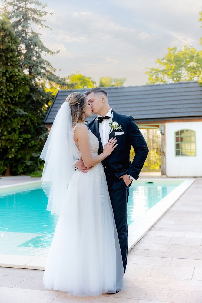 Newlyweds share a romantic moment by a pool with a white house in the background.