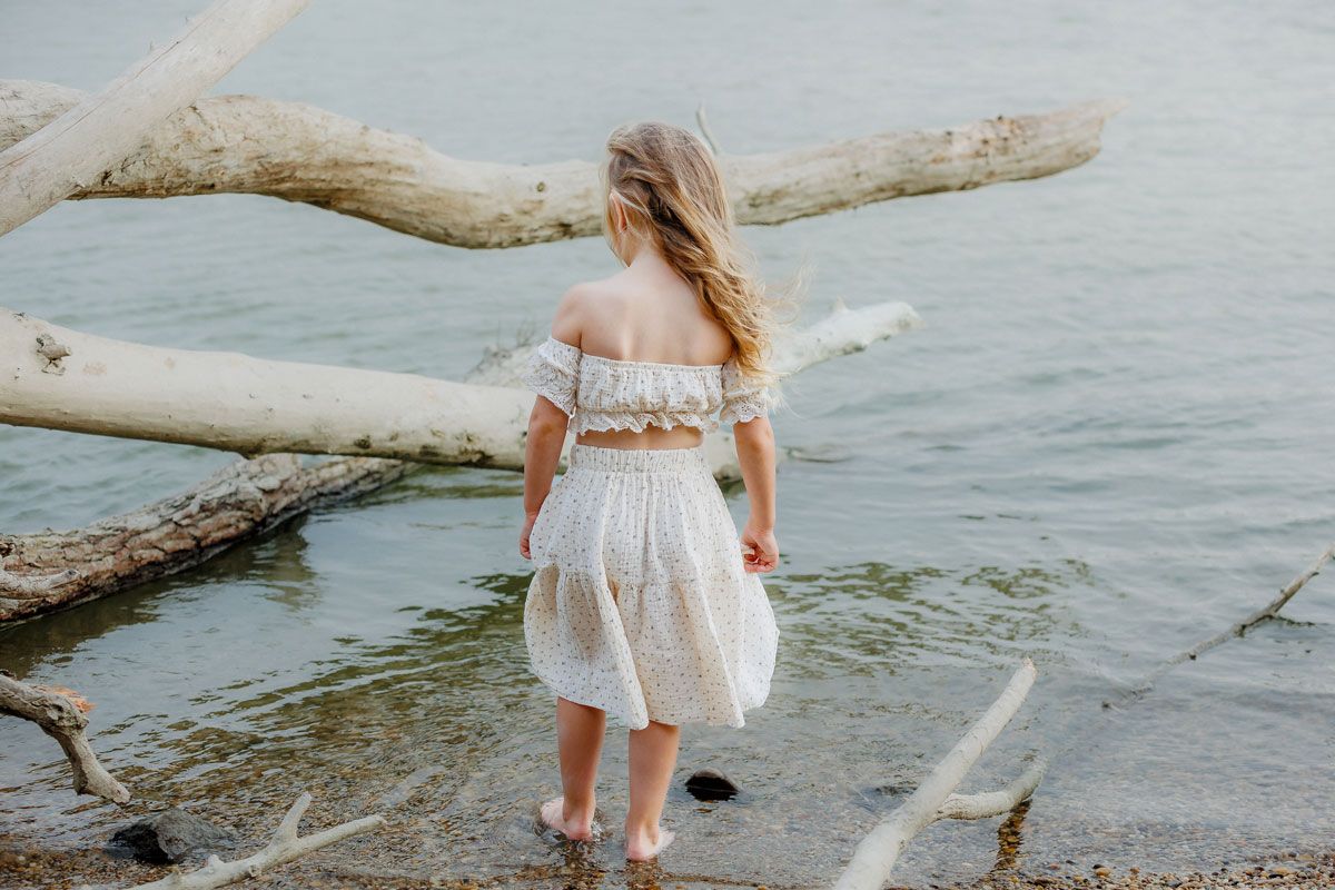 Young girl in white off-shoulder crop top and skirt wading in shallow water near driftwood on a rocky shore.