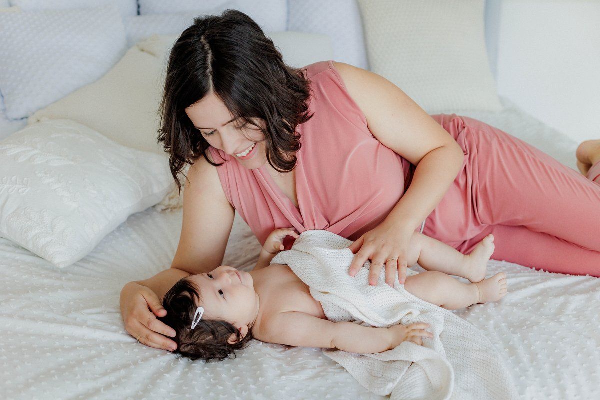 A smiling mother in a pink dress lying on a bed, lovingly gazing at her newborn baby.