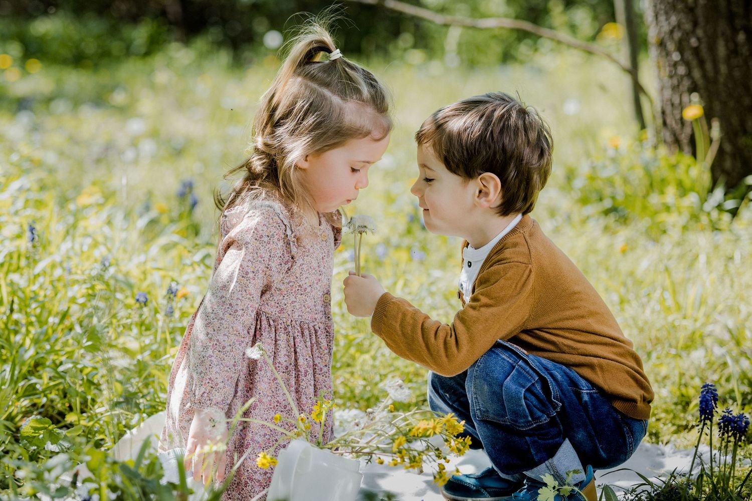 Two young children sit in a flower field, a girl in a floral dress and a boy in mustard yellow sharing a dandelion.