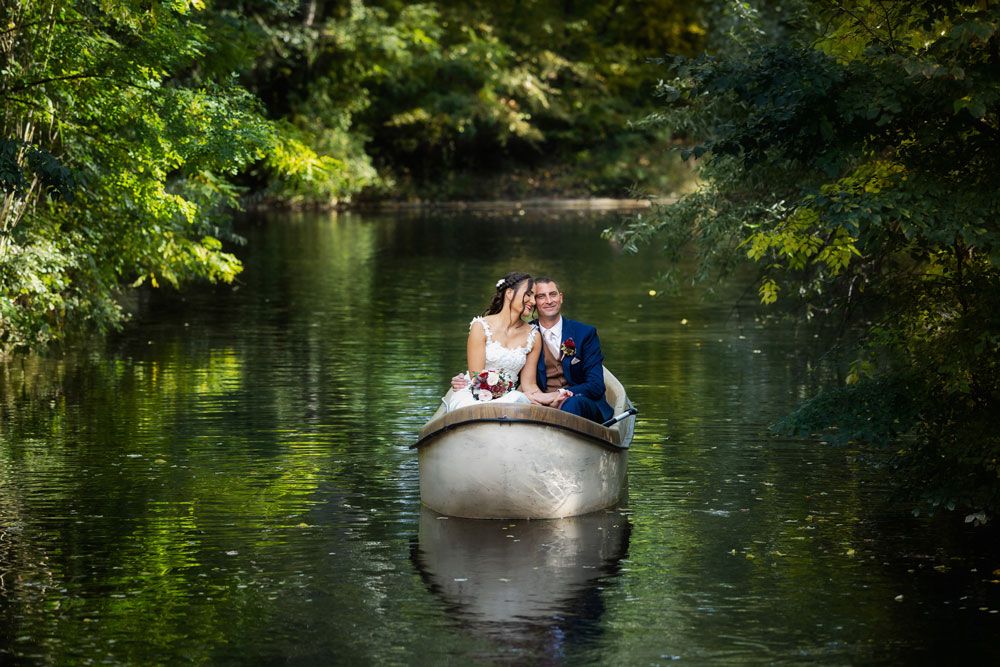 A couple in formal attire sits together in a small boat on a serene, tree-lined river with green reflections.