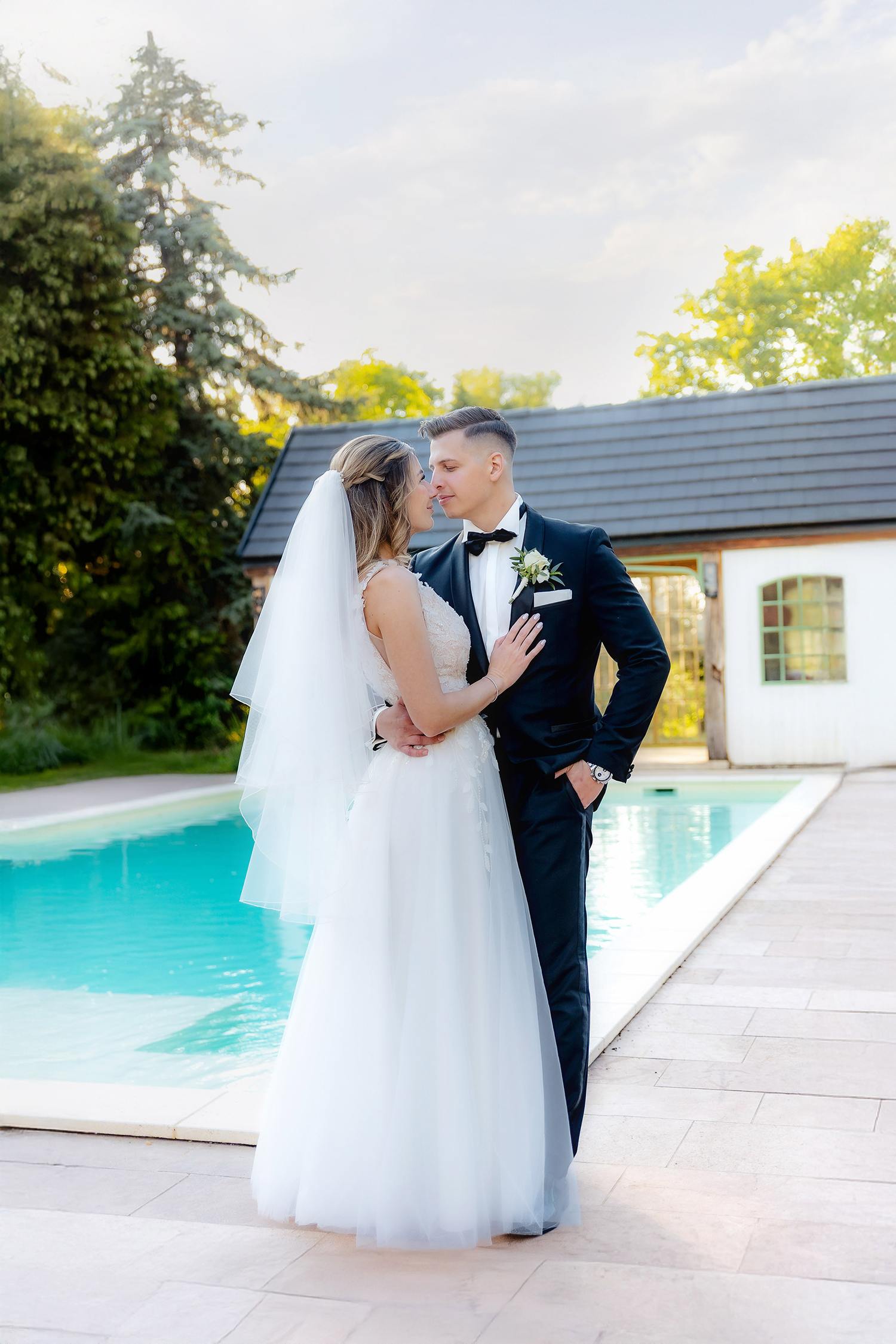 Bride and groom share a romantic kiss by a pool, dressed in a white gown and black tuxedo on their wedding day.