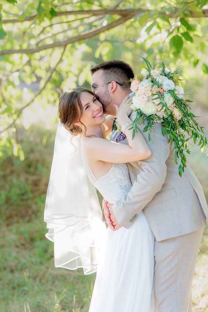A couple in wedding attire embraces under sunlit green trees in a romantic outdoor setting.