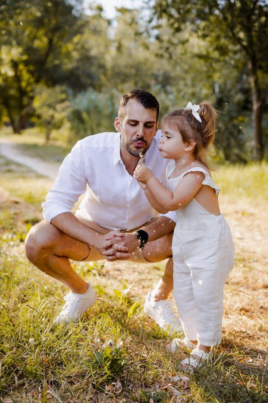 Father and toddler daughter in white outfits blowing dandelion together in sunny park setting.