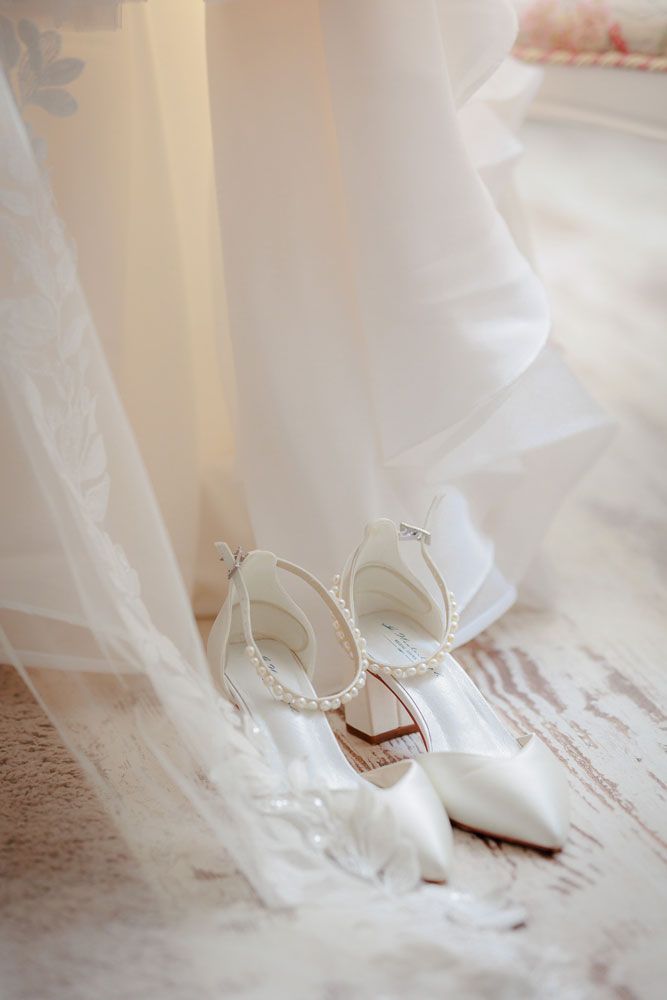 White bridal shoes peek out from beneath delicate tulle fabric on a rustic wooden floor.