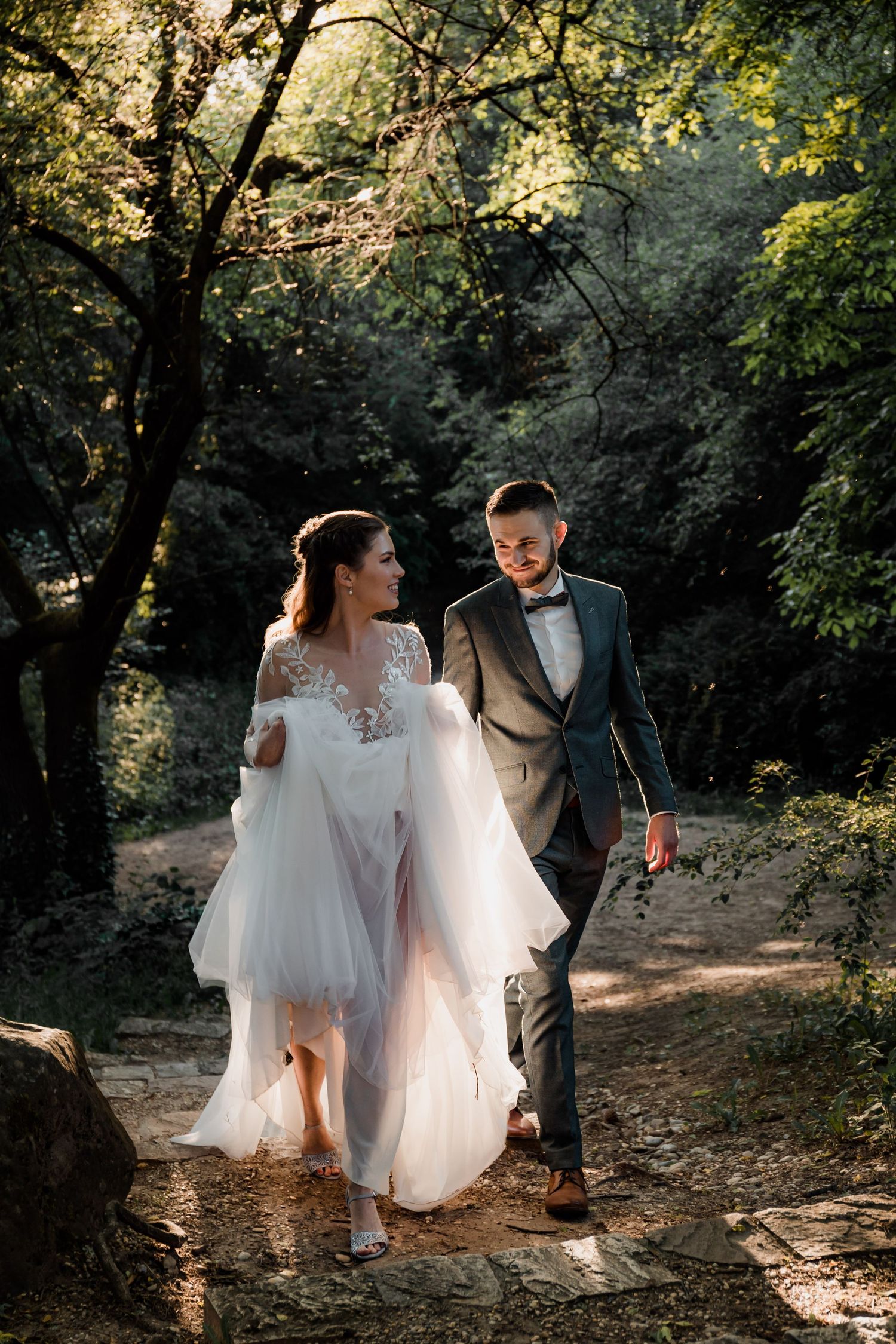 Bride and groom walk hand in hand through a sunlit forest path, she in a flowing white gown, he in a gray suit.