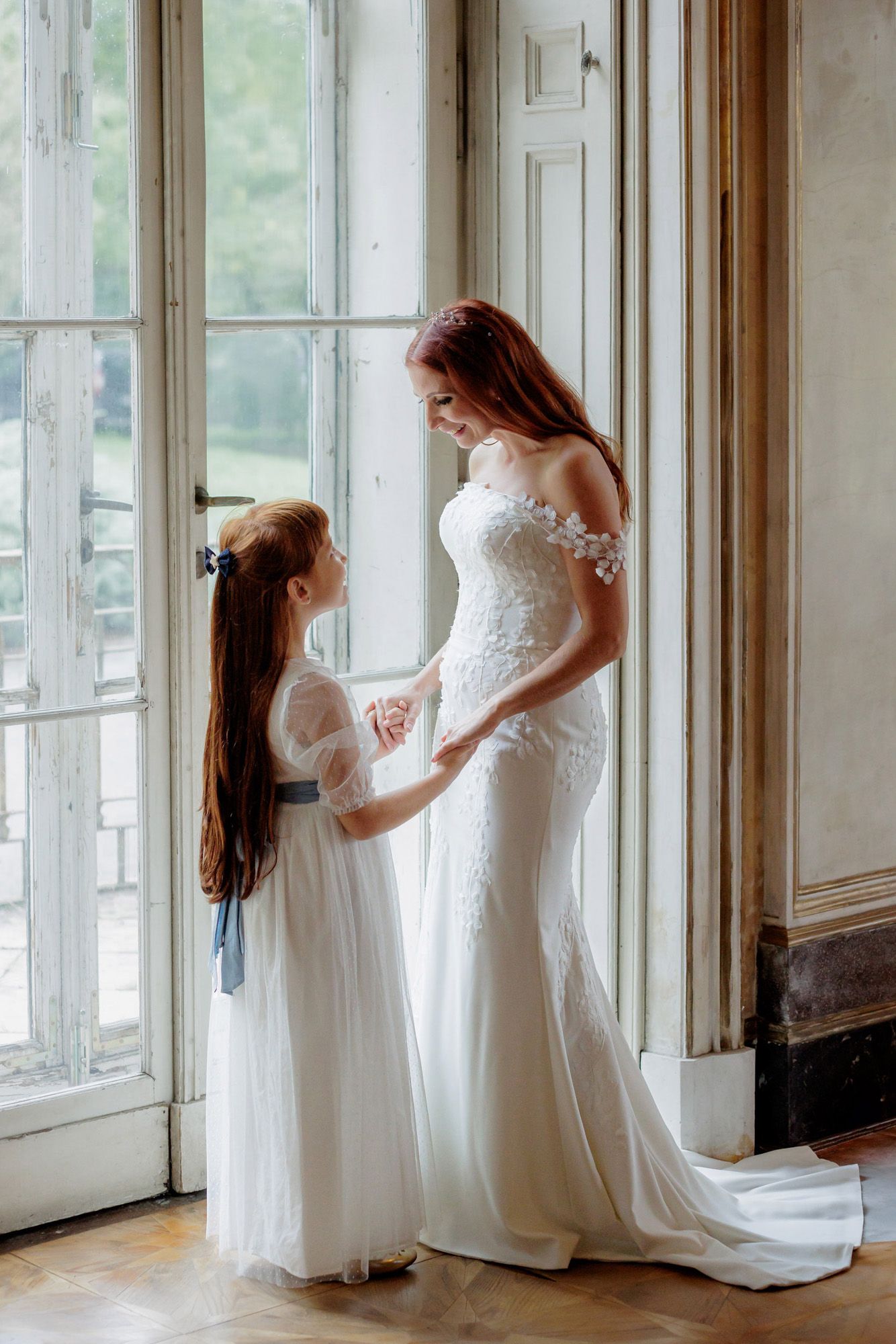 Bride in lace gown holds hands with flower girl in white dress by elegant French doors.