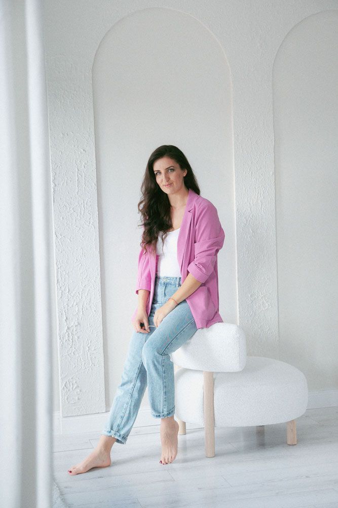 Woman in pink cardigan and denim overalls sits in white minimalist space creating artistic movement series.