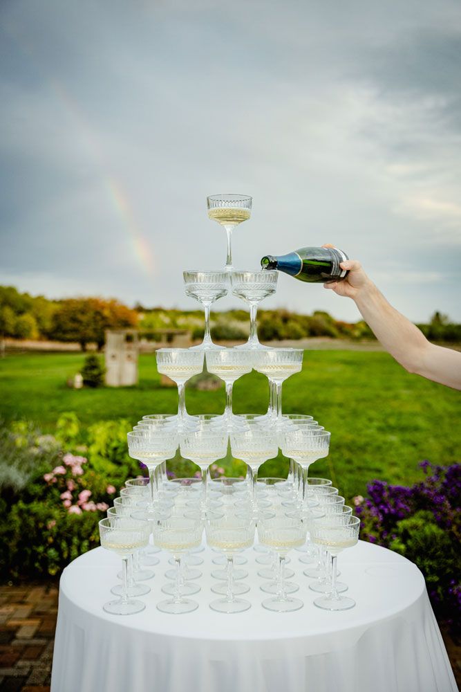 A champagne tower with crystal coupe glasses stacked in pyramid formation on a white tablecloth outdoors at sunset.