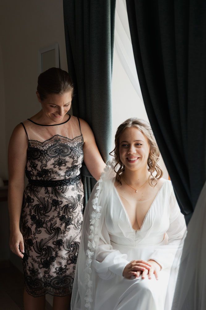 Two women stand by a window, one wearing a black lace dress and the other in a white bridal robe.