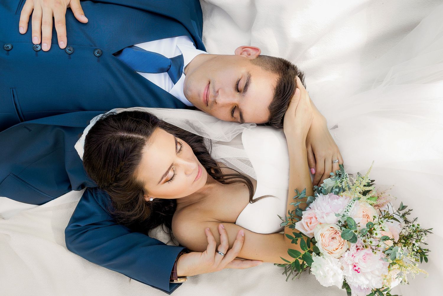 Bride and groom in wedding attire lying together, bride holding a pastel bouquet, eyes closed in a peaceful embrace.