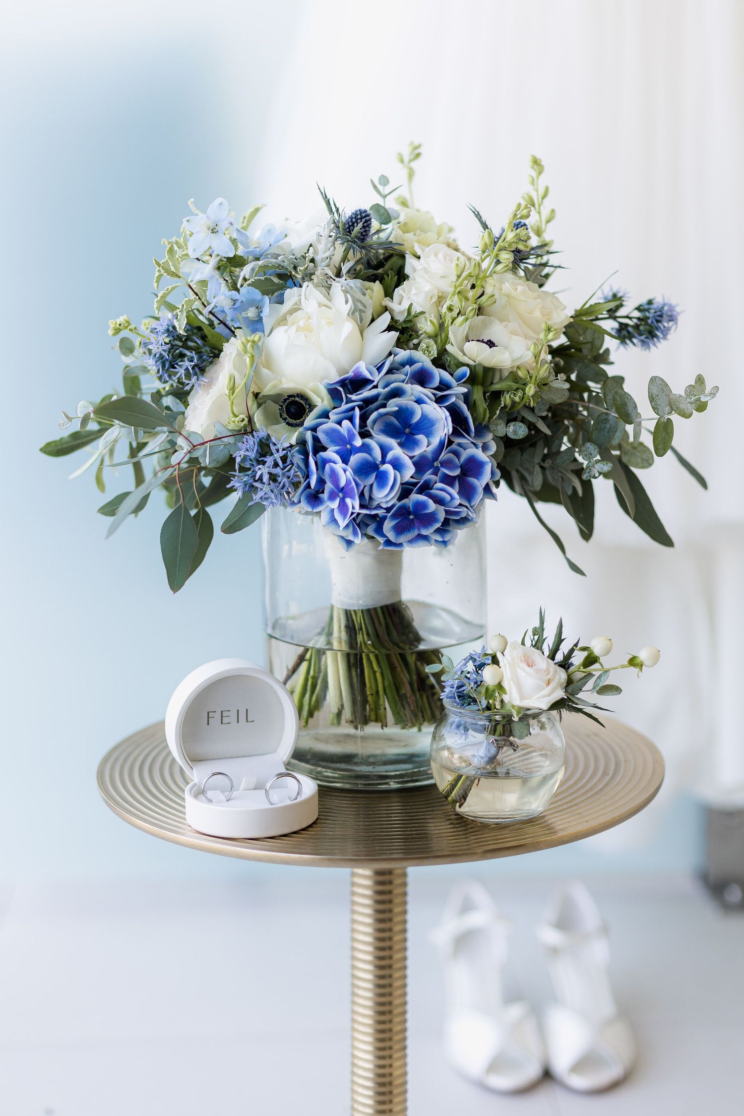 Blue and white wedding bouquet with hydrangeas in glass vase, ring box, boutonniere, and white heels on gold table.