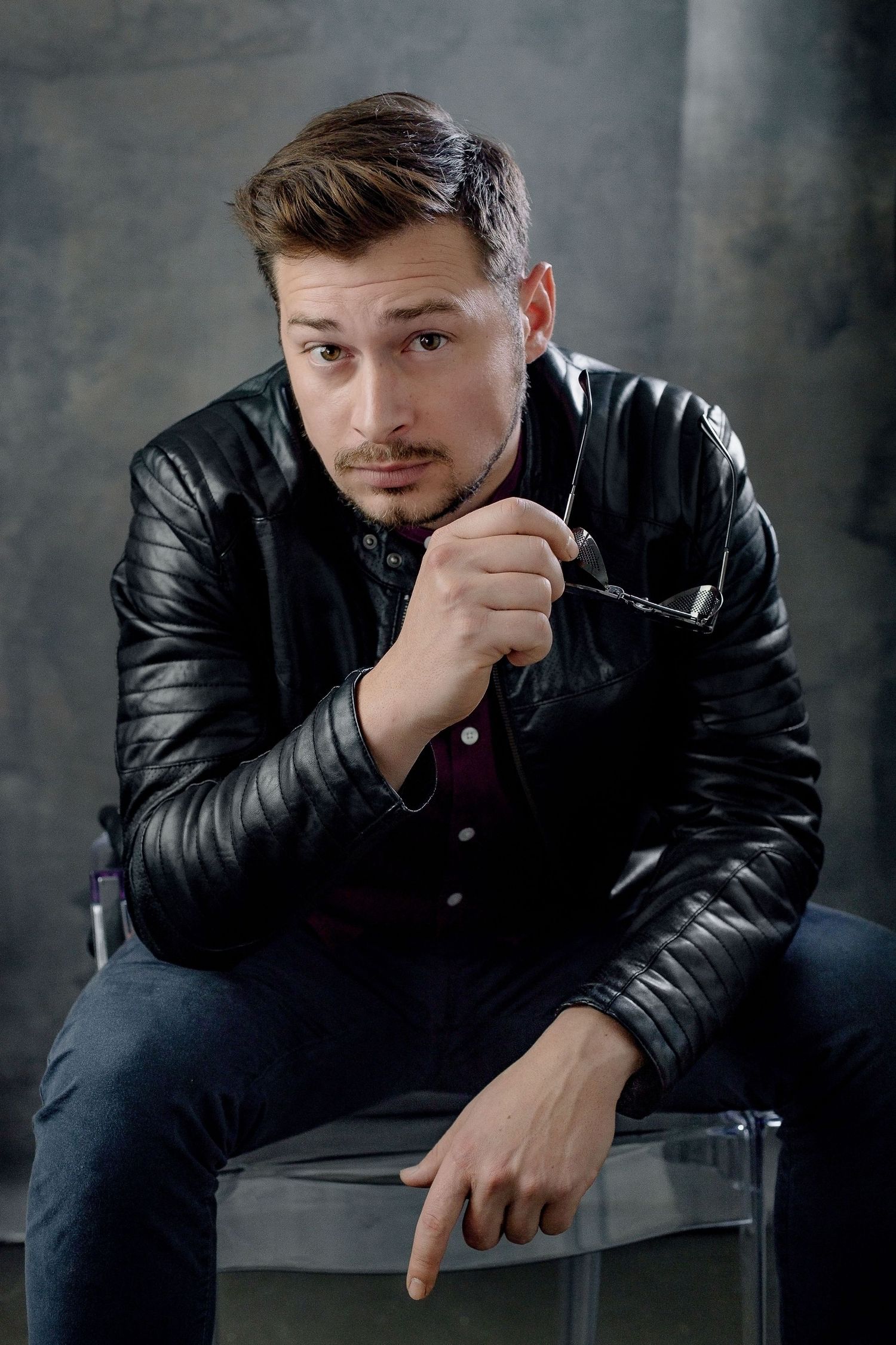 Man in black leather jacket sitting on chair, holding glasses, looking at camera with confident expression.