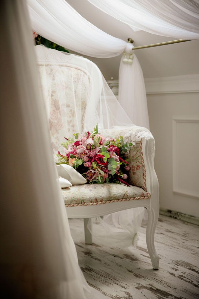 Elegant white chair with pink floral cushions and flowing sheer curtains in a bright, romantic setting.