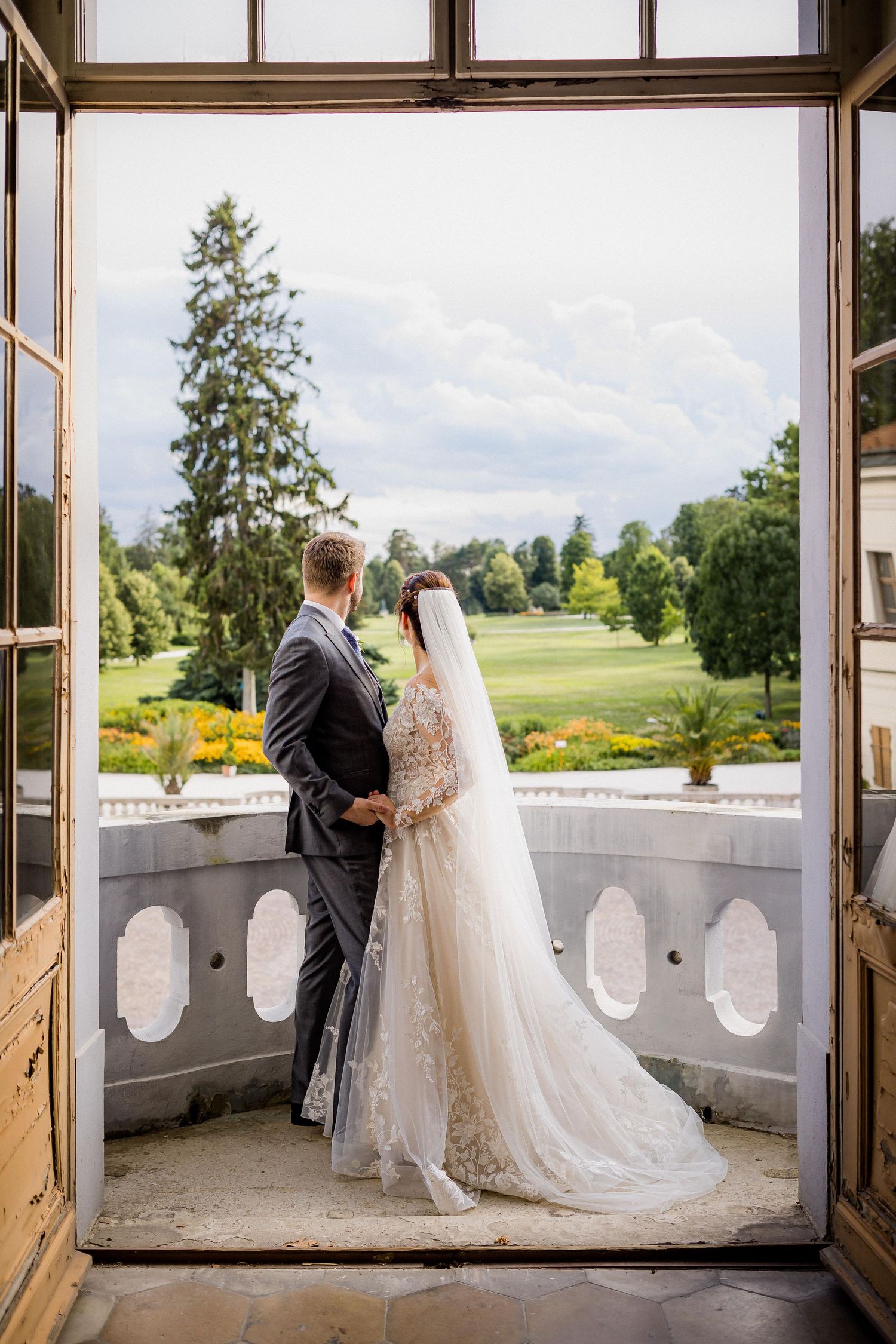 Bride and groom stand on elegant balcony overlooking lush green garden, bride wearing lace gown with long veil.