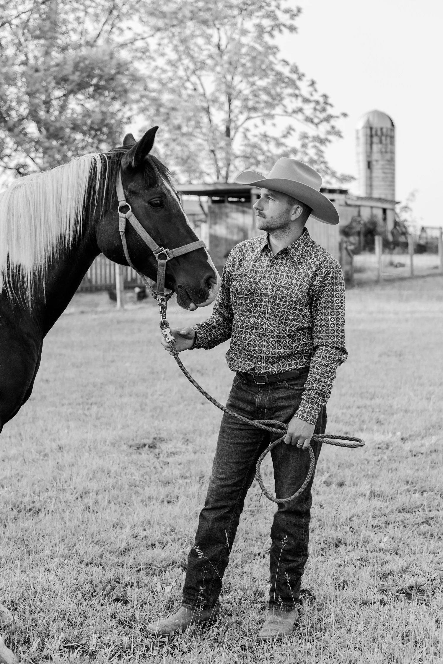 A person in western attire stands beside a black horse in a grassy field.