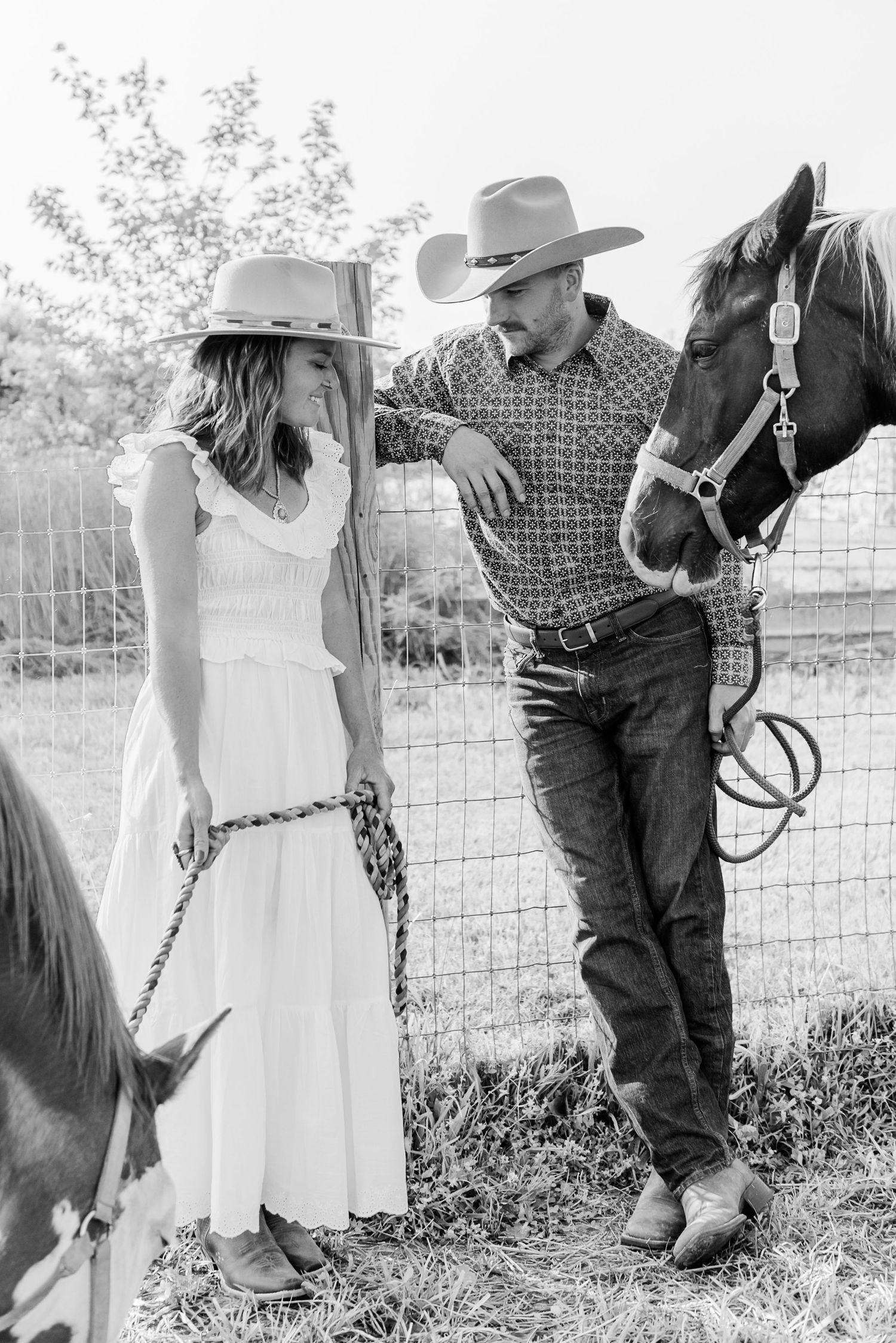 A couple in western attire stands by a fence with their horses on a sunny day in the countryside.