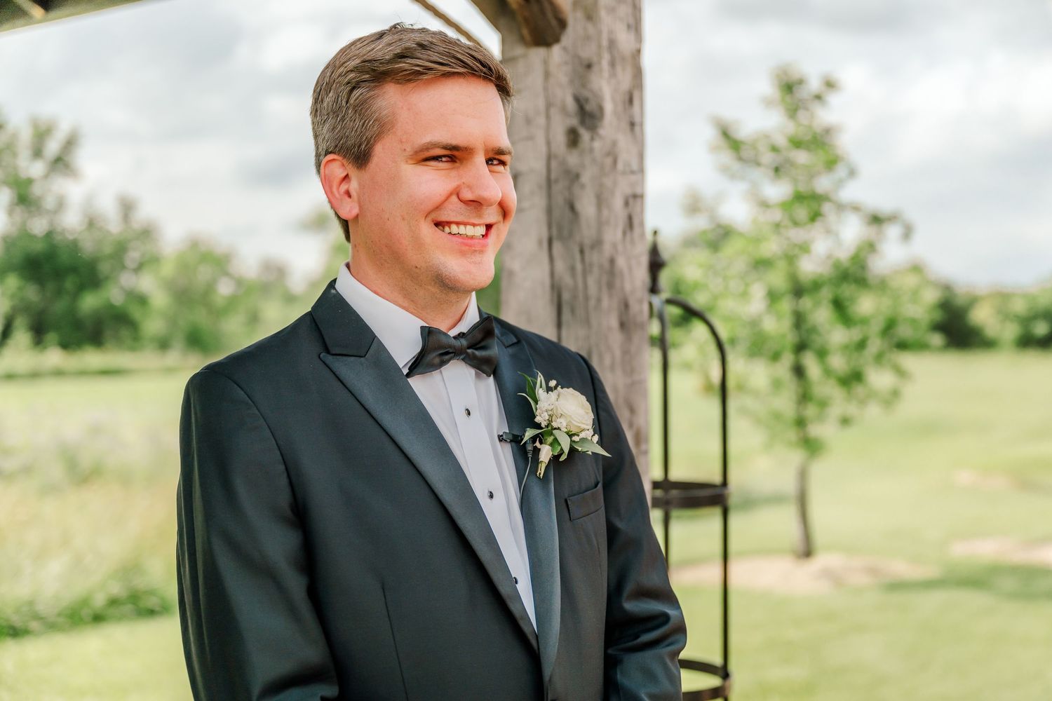 A groom in a classic black tuxedo and gray tie smiles while standing outdoors near trees on a sunny day.