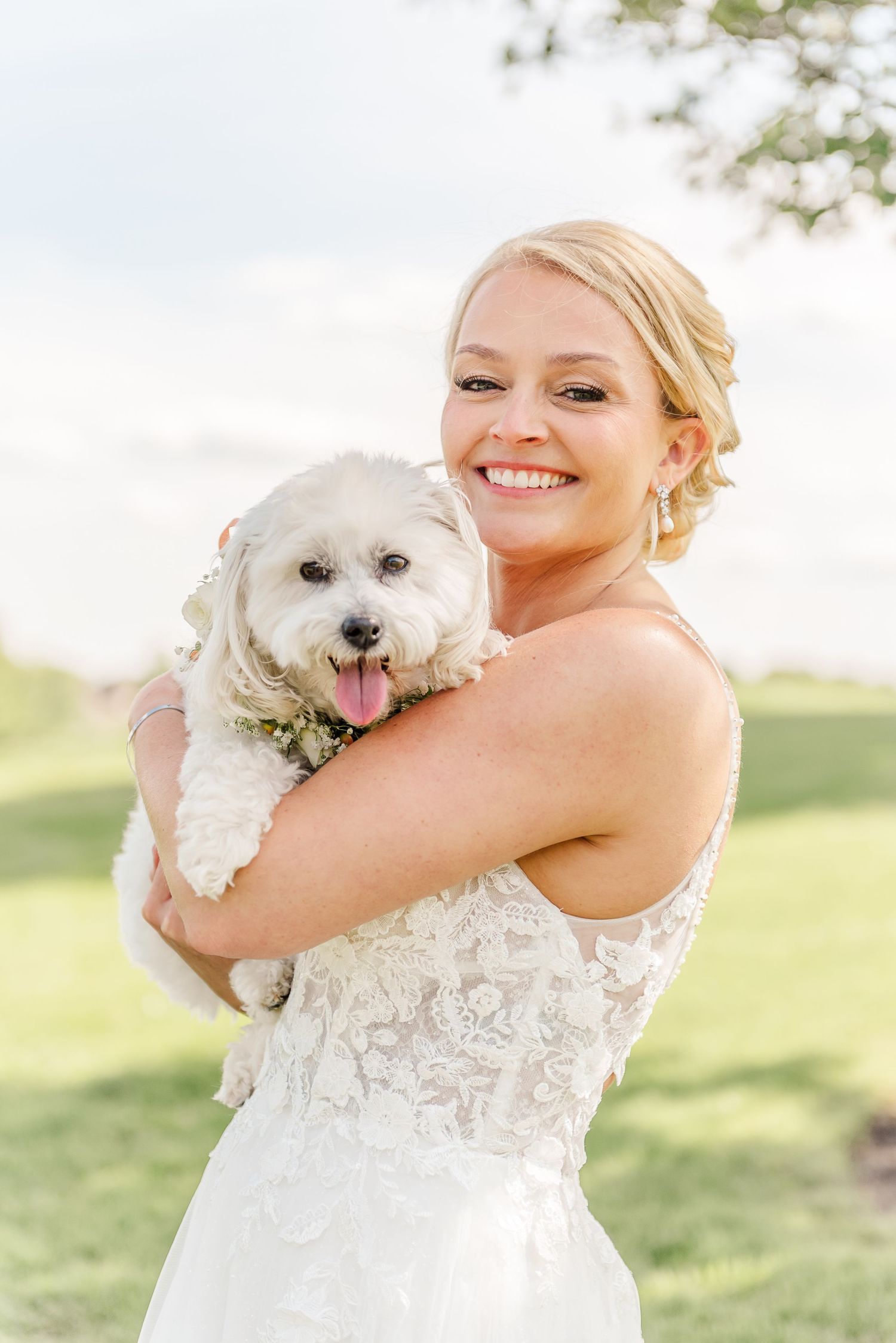 A bride in a lace wedding dress holds and cuddles with a small white fluffy dog outdoors on a sunny day.