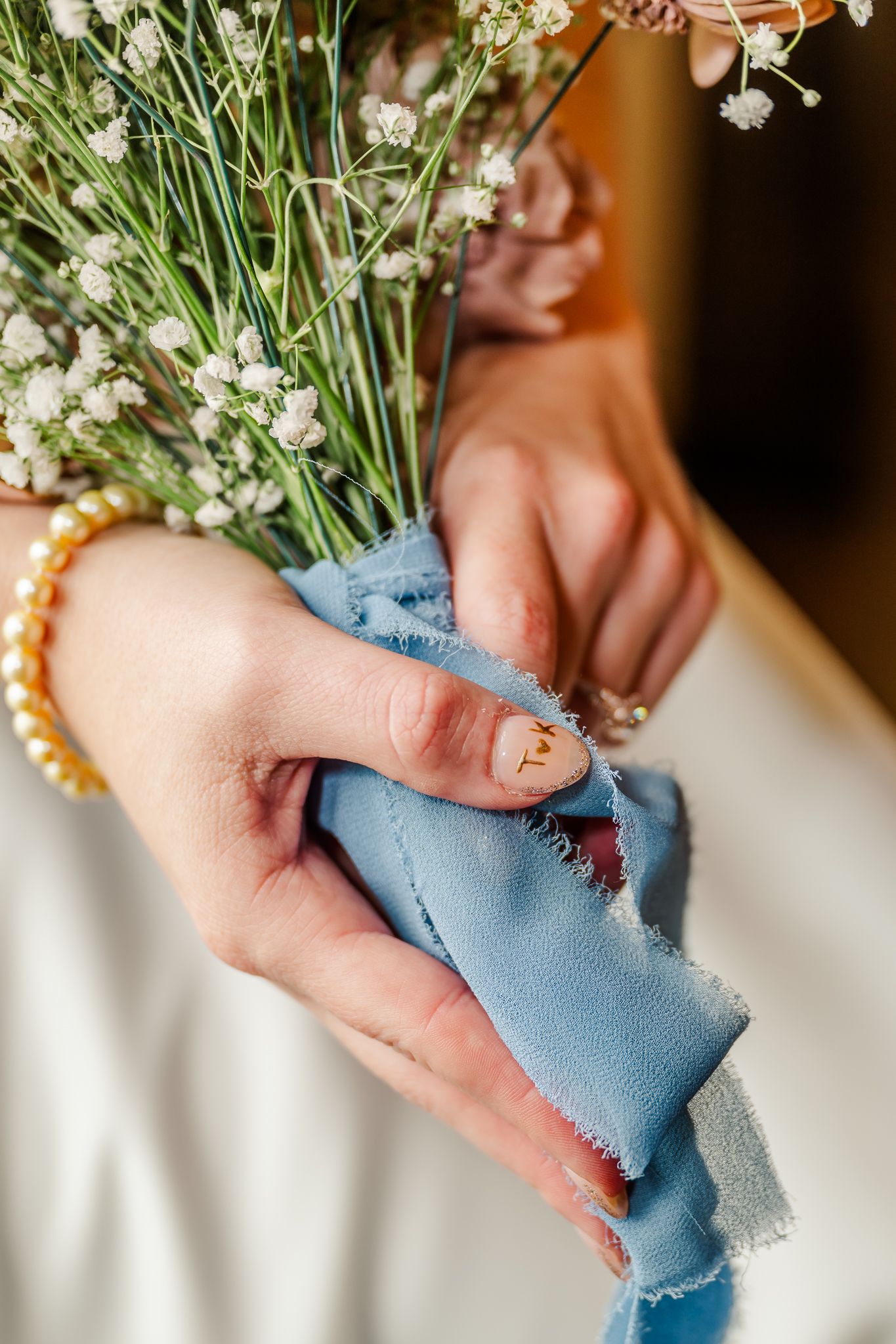 Close-up of hands holding a white bouquet tied with a blue ribbon, wearing pearl bracelet and ring. The Old Barns at Dry Run Farms