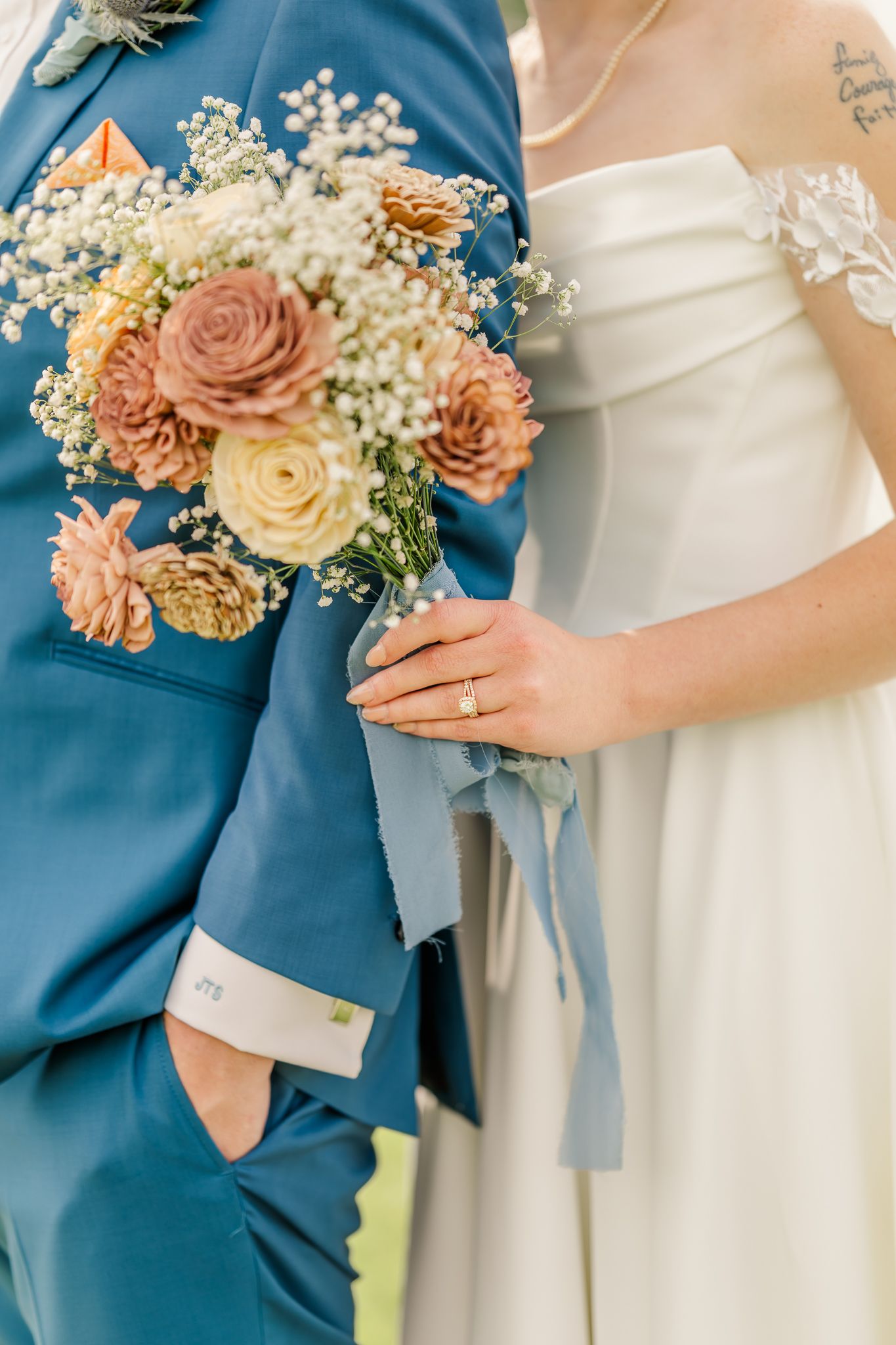 Bride holding a rustic bouquet with dried flowers and baby's breath, standing beside groom in blue suit.