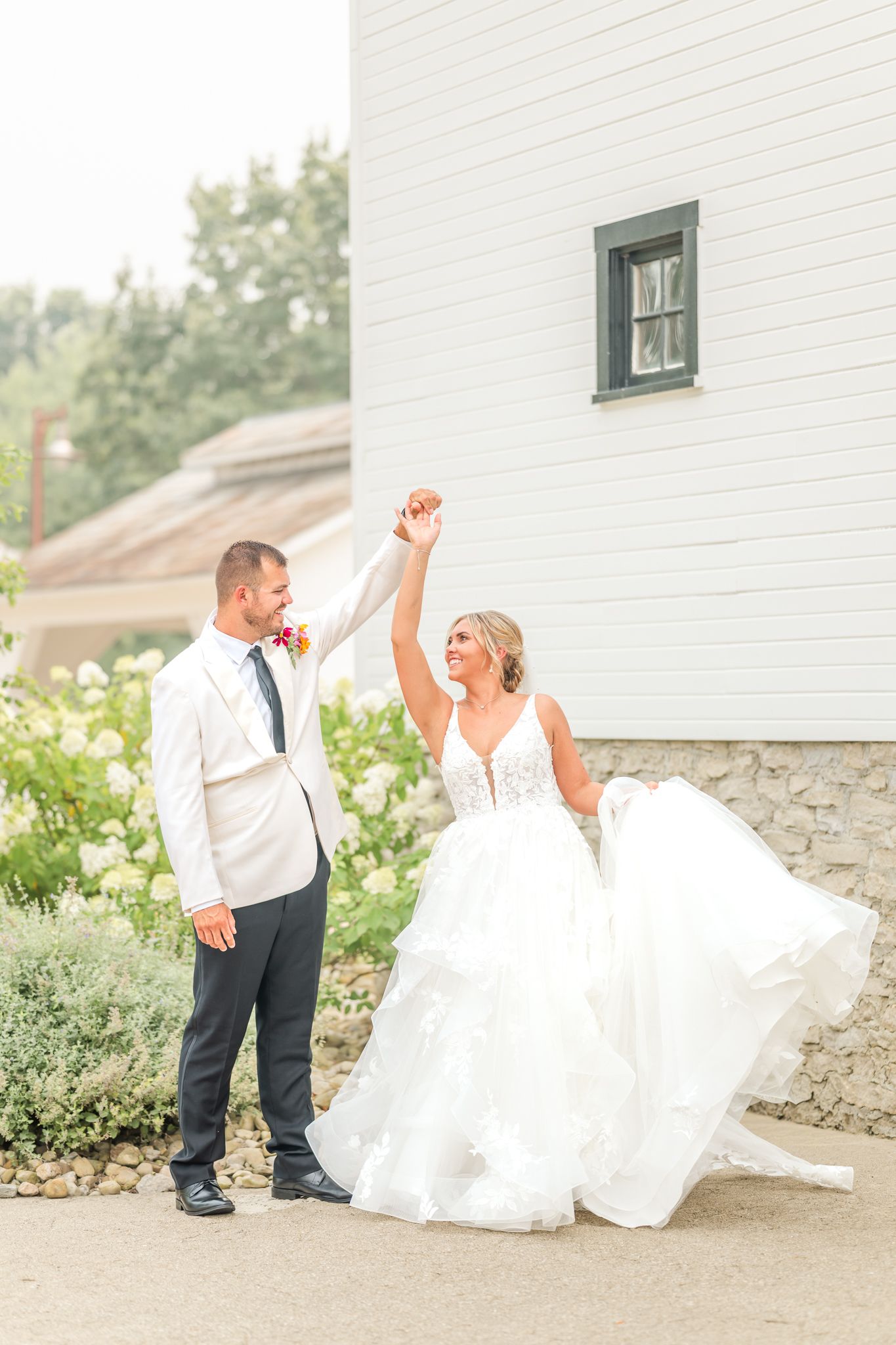 Bride in flowing white gown twirls as groom in white jacket holds her hand outside a white barn. The Crawford Barn in Frazeysburg, Ohio.