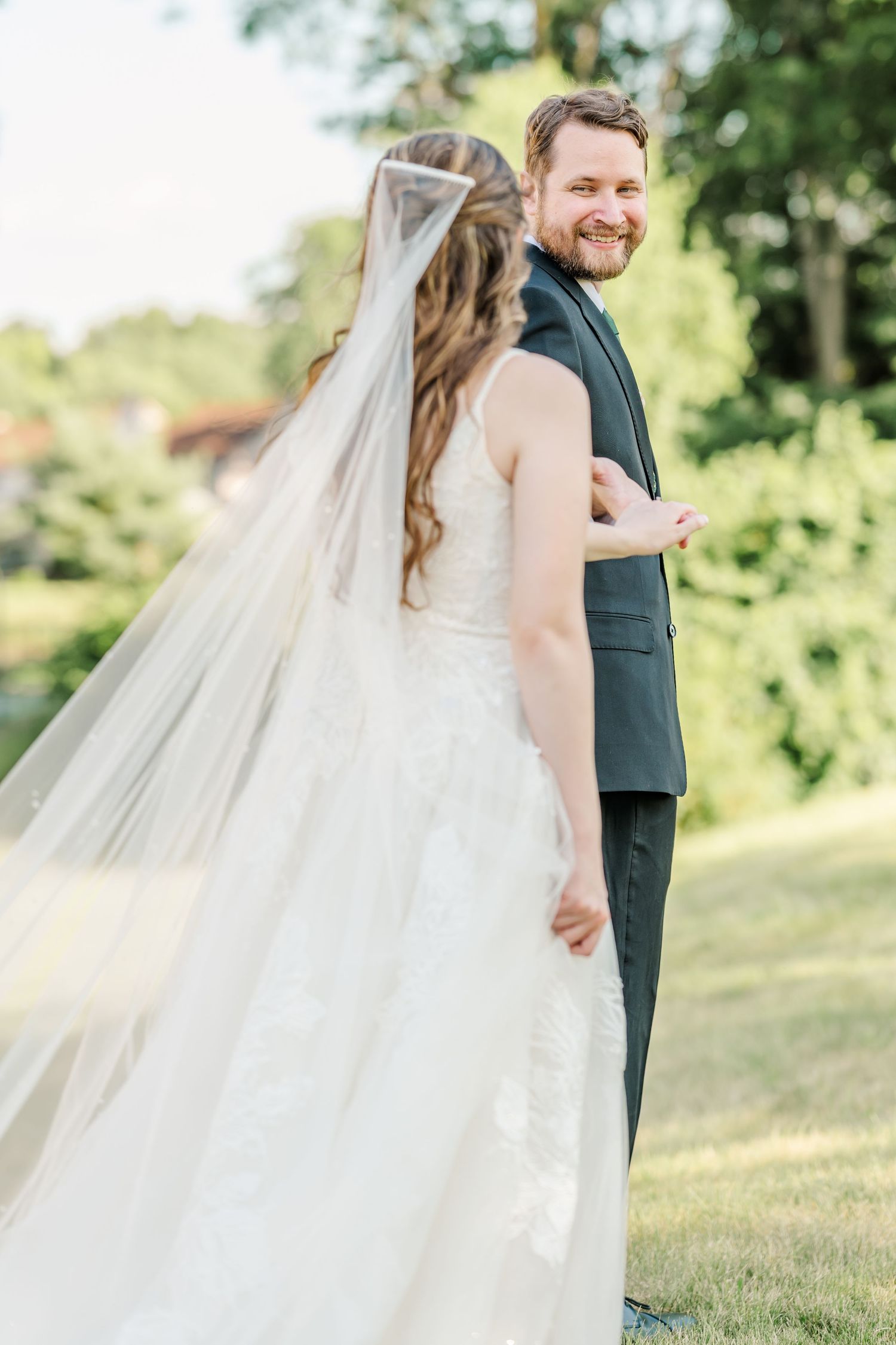 Couple in wedding attire standing back-to-back outdoors with flowing veil in sunlight.