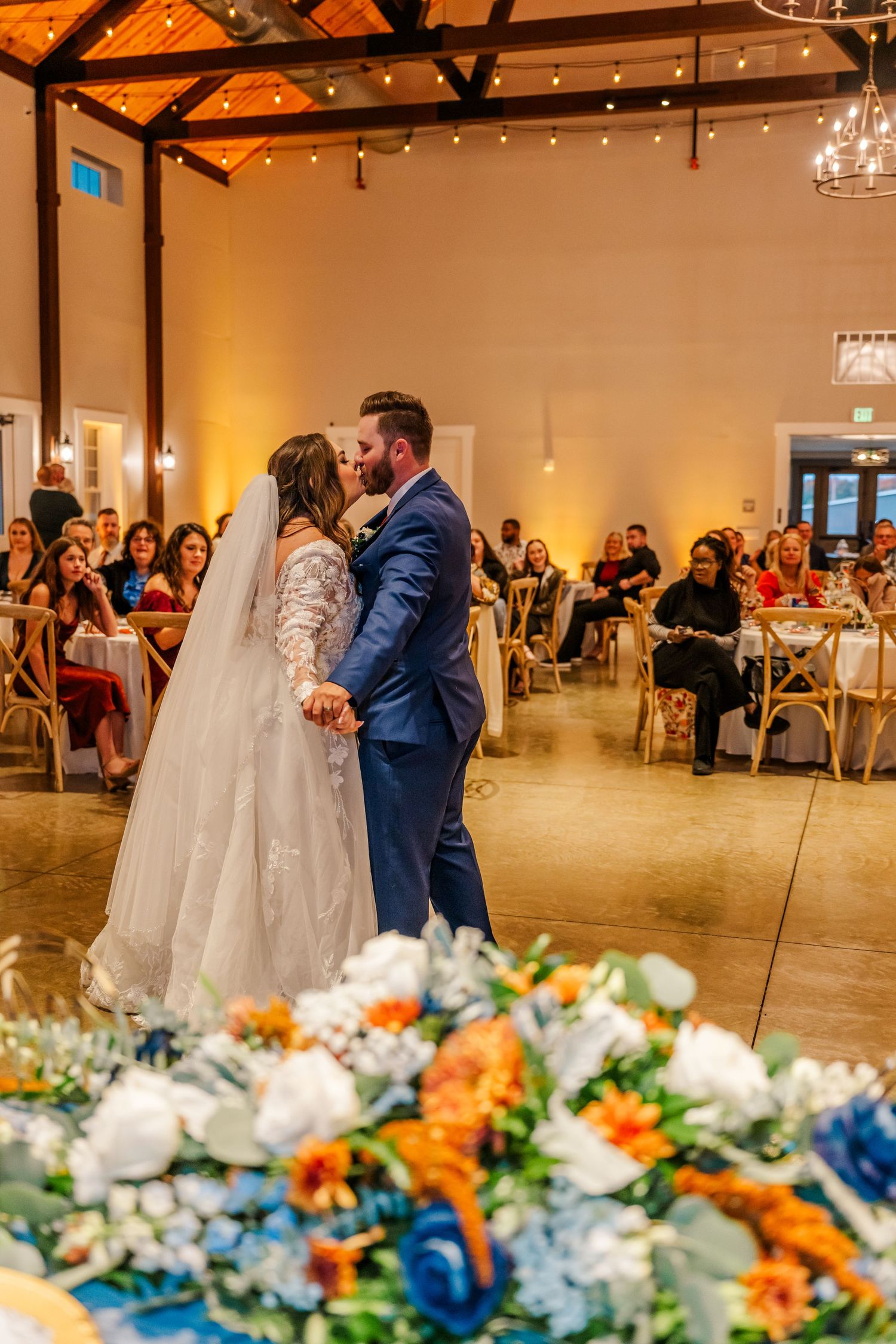 Couple shares their first dance at a wedding reception with orange and blue floral decorations in the foreground.