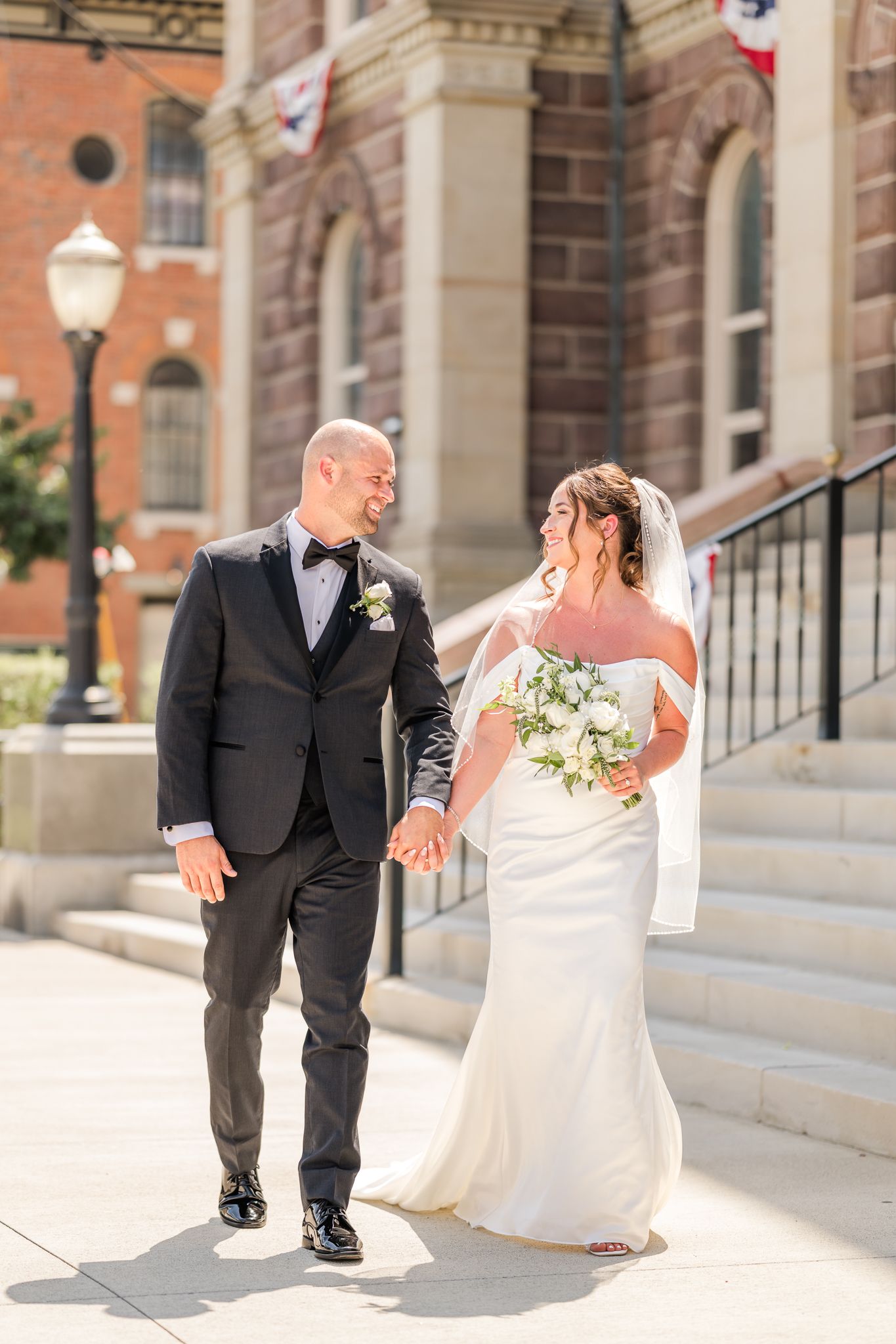 A bride & groom walk hand-in-hand outside a historic brick building, smiling at each other on their wedding day. Pickaway county courthouse