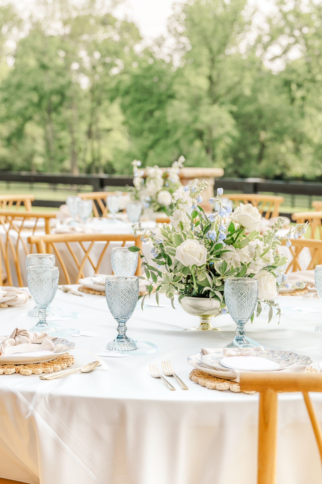 Elegant outdoor wedding reception table with white floral centerpiece, blue goblets, gold chargers, and white linen tablecloth Canopy Creek