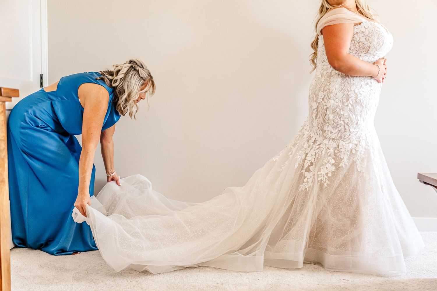 Bridesmaids in royal blue dresses help adjust a bride's flowing white wedding gown train in a bright room.