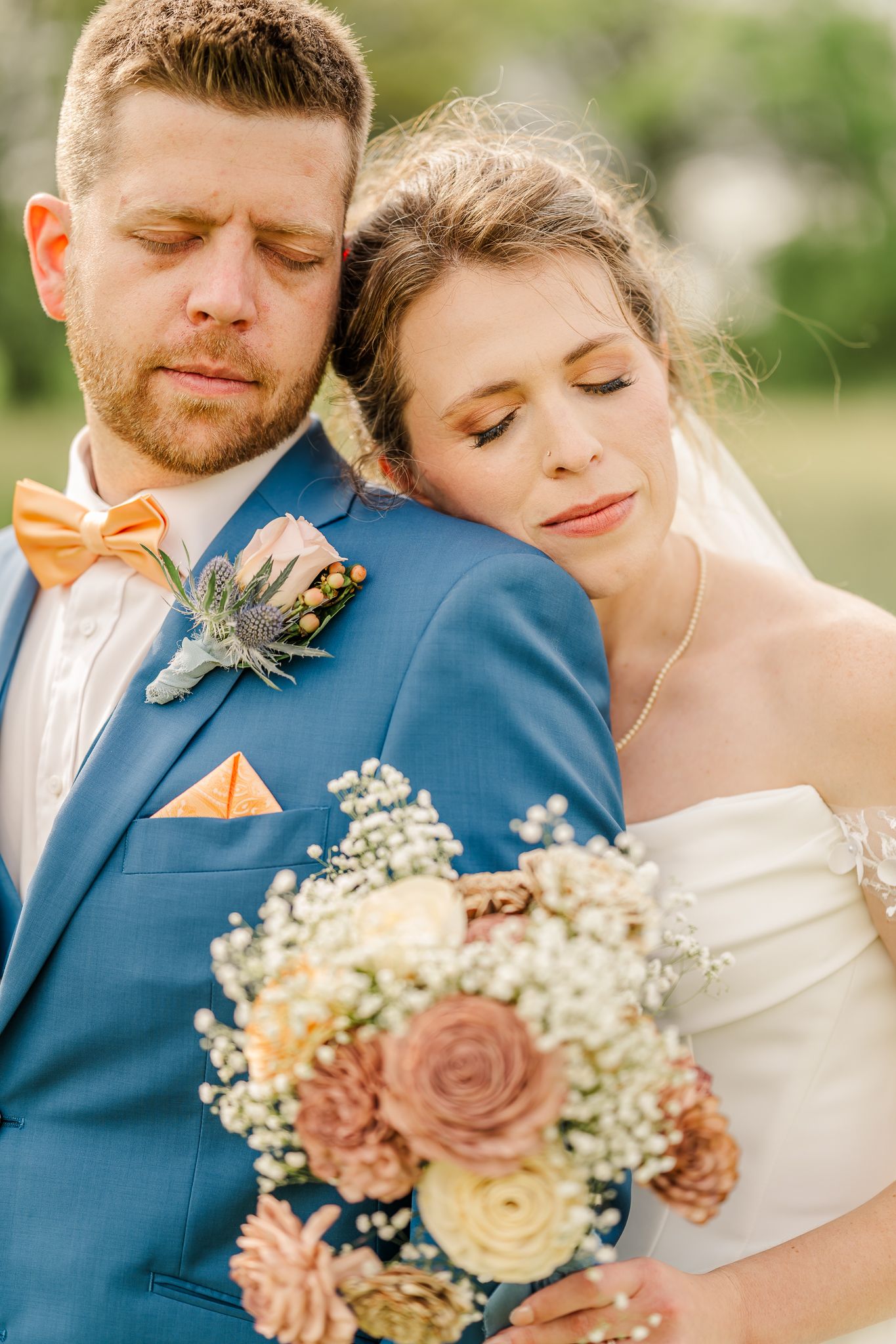 Bride rests head on groom's shoulder, both eyes closed, as she holds a pastel bouquet, groom in blue suit with orange bow tie.