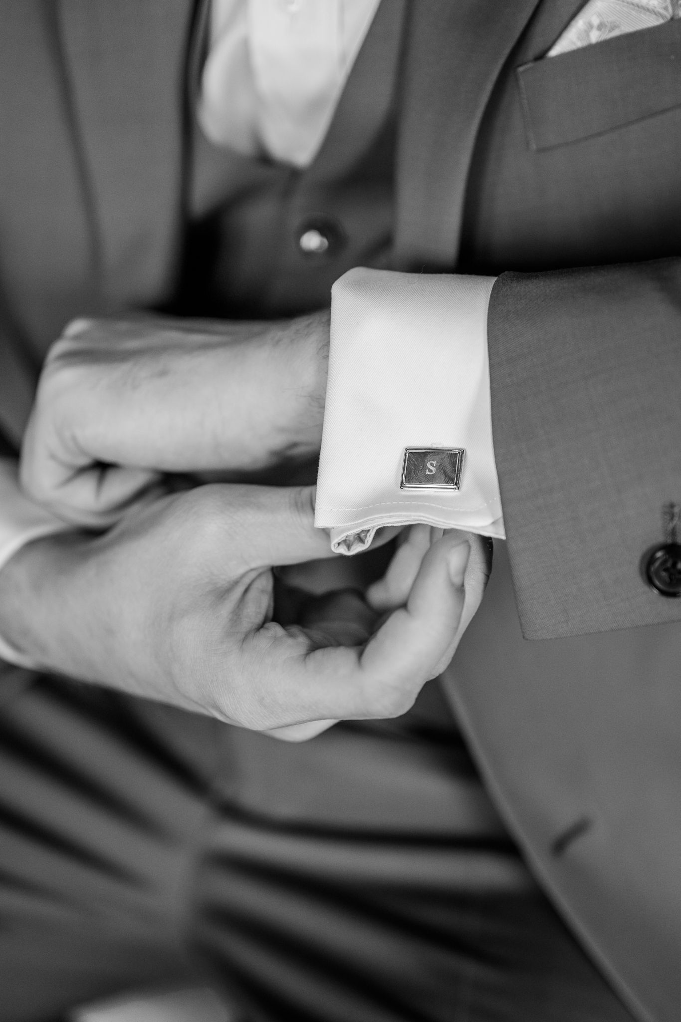 Black and white photo of a groom fastening monogrammed cufflinks on his suit sleeve before the wedding.