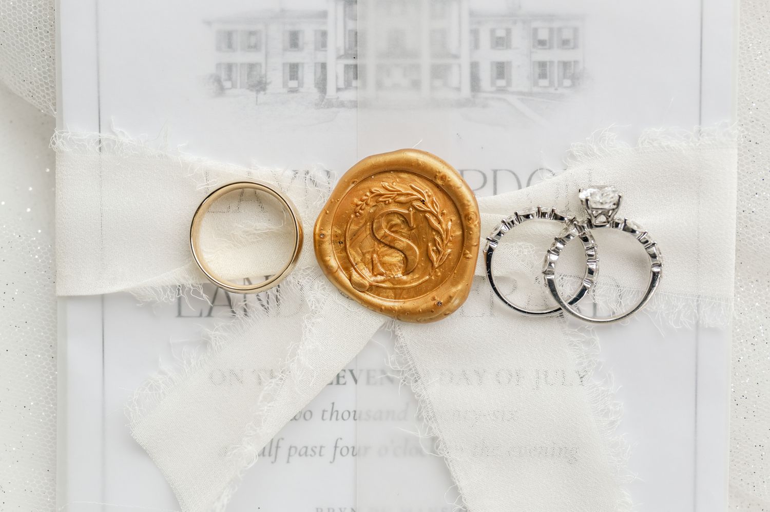 Gold wax seal monogram with white ribbon beside wedding rings on elegant invitation suite.