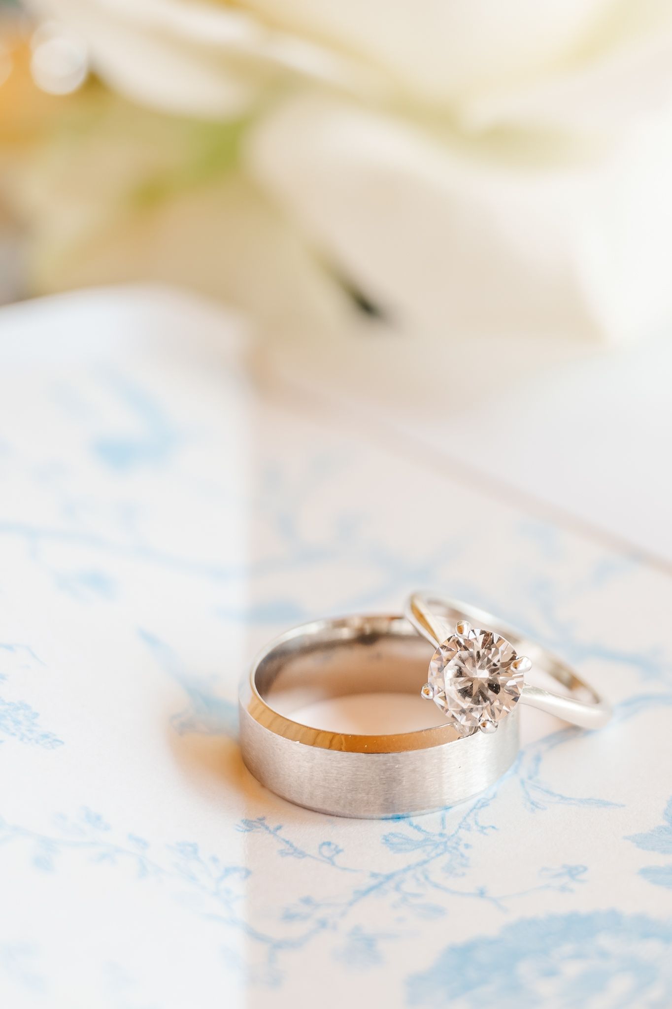 Wedding rings set on blue floral fabric, featuring a diamond solitaire engagement ring and brushed platinum band.