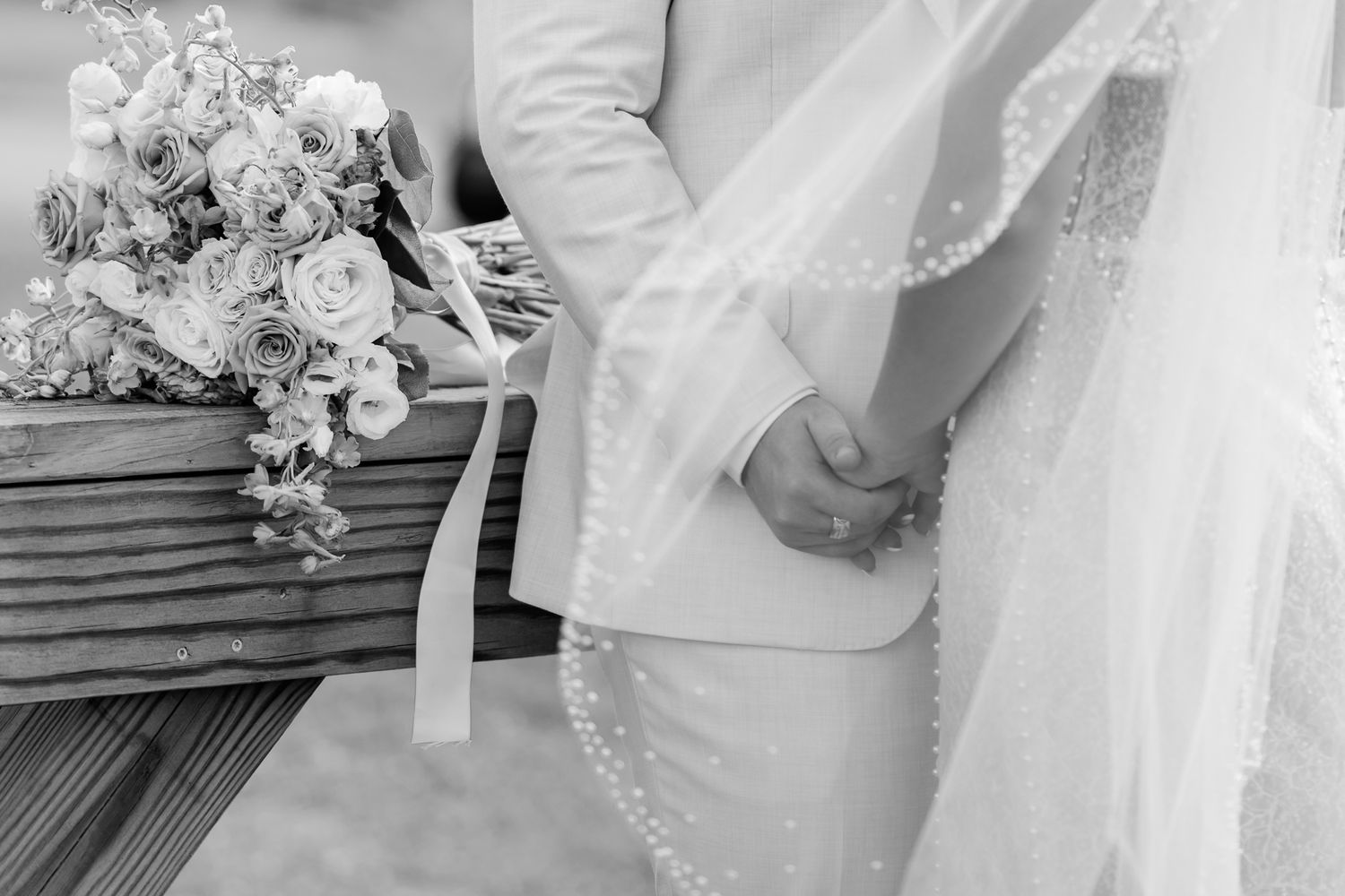 Black and white wedding photo of bride and groom holding hands near a floral bouquet with ribbon.
