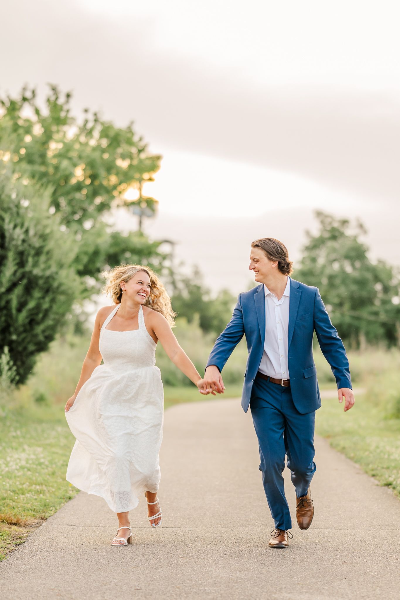 A joyful couple holds hands and walks down a path, the woman in a white dress and man in a blue suit, surrounded by greenery. Scioto Audubon