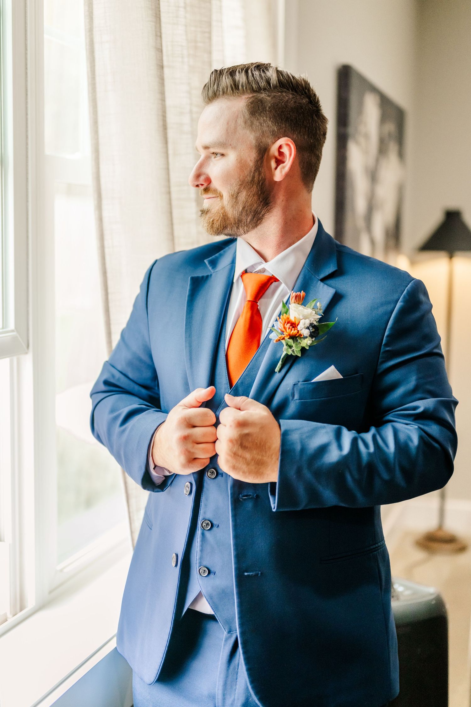 Groom in blue three-piece suit with orange tie adjusts his jacket by window.