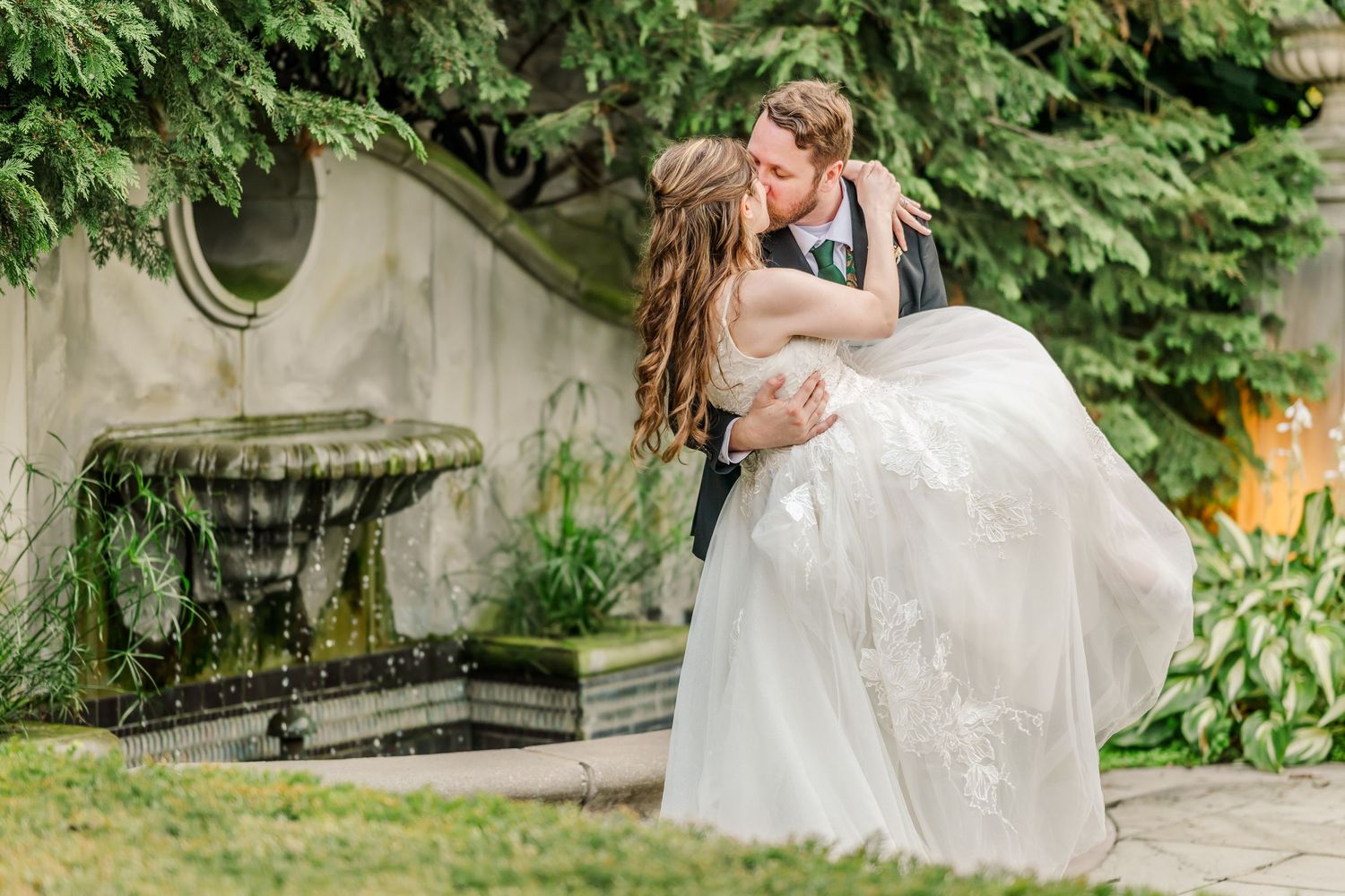 A romantic wedding couple embraces in a garden setting with an ornate stone fountain and lush greenery in the background.